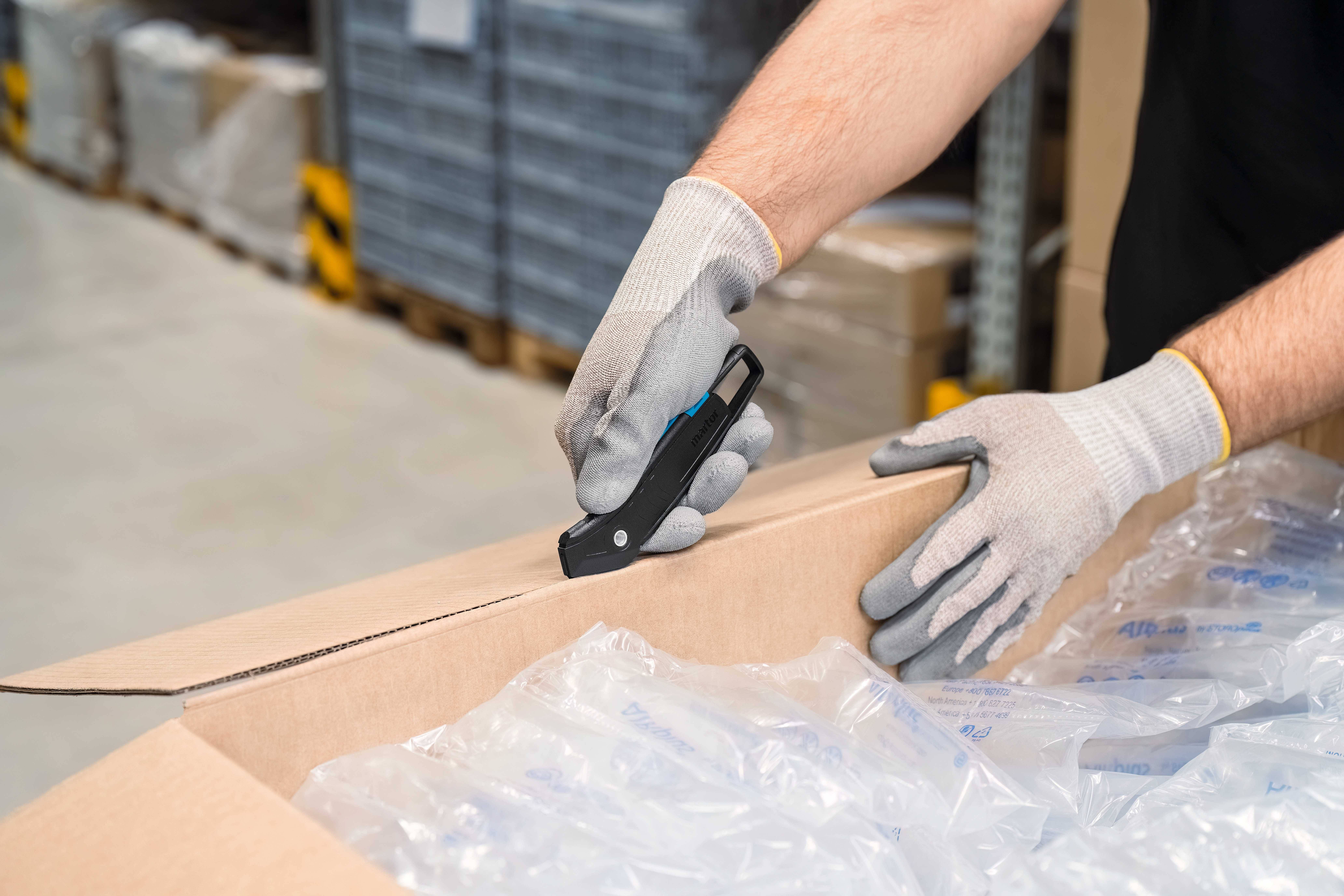 A person wearing grey gloves is cutting a cardboard box with a utility knife in a warehouse. Shelves are visible in the background.