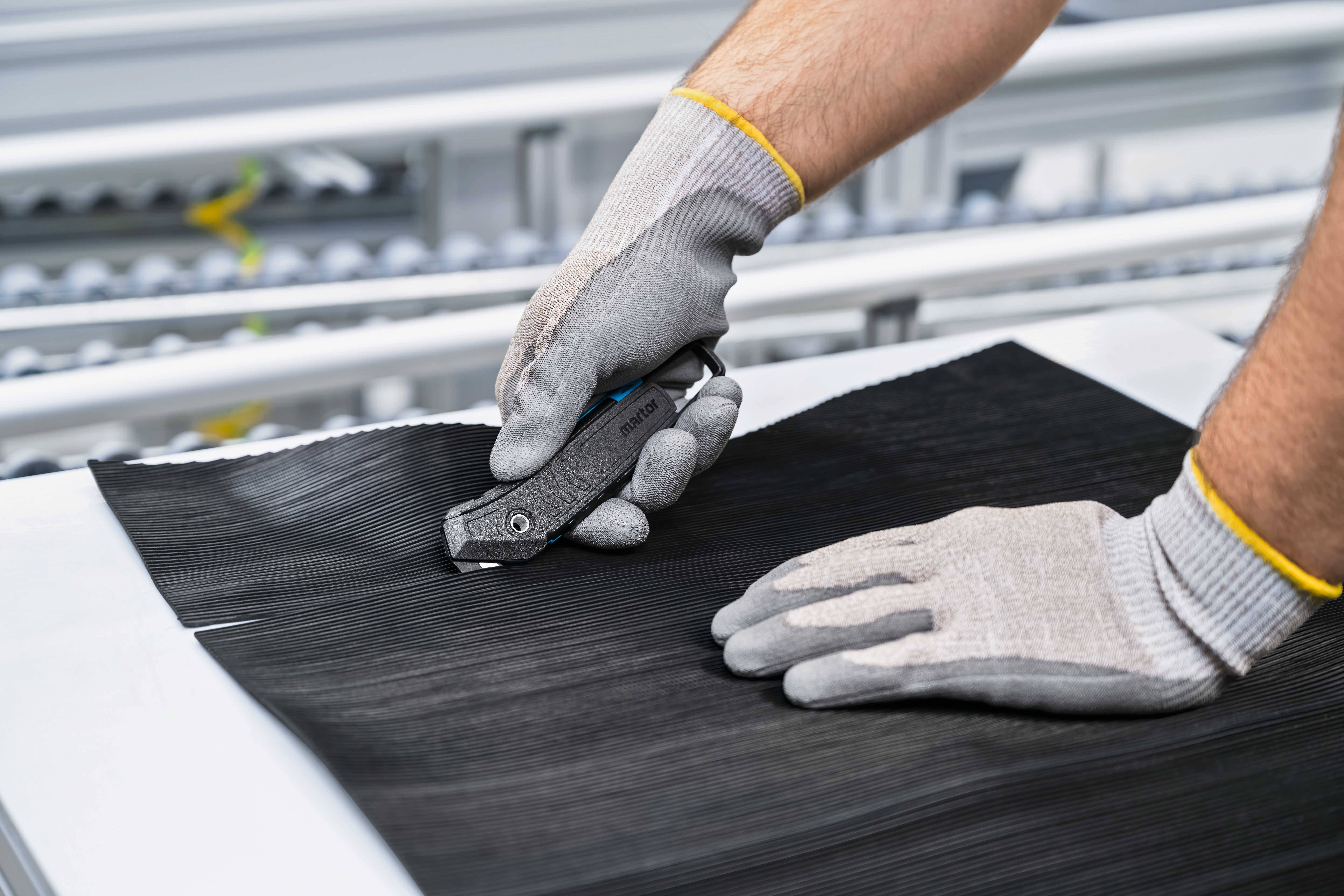 A person is cutting a black material with a grey knife on a table. They are wearing grey protective gloves.