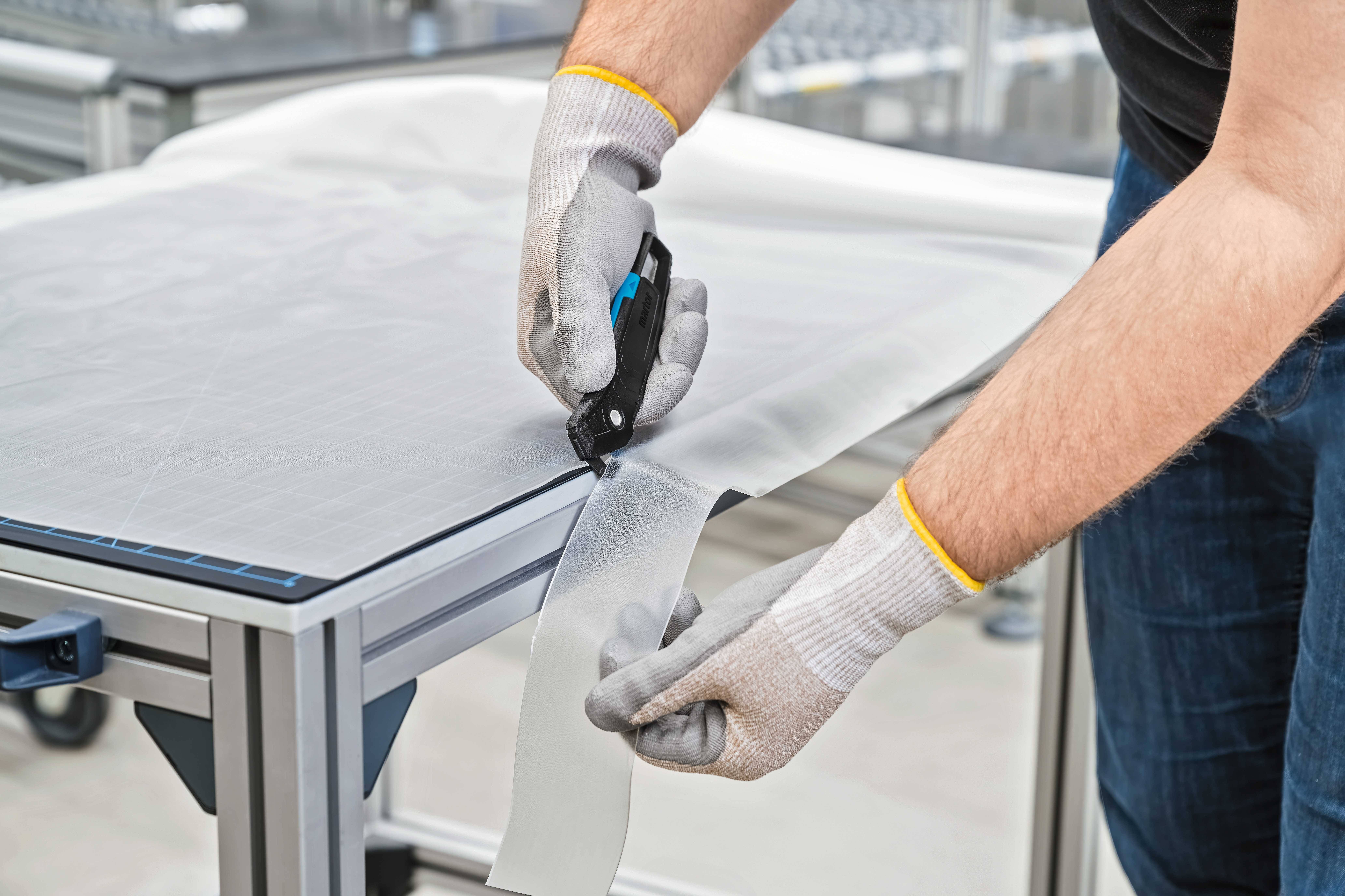 A man is cutting a piece of tape from a workbench using a safety knife. He is wearing protective gloves.