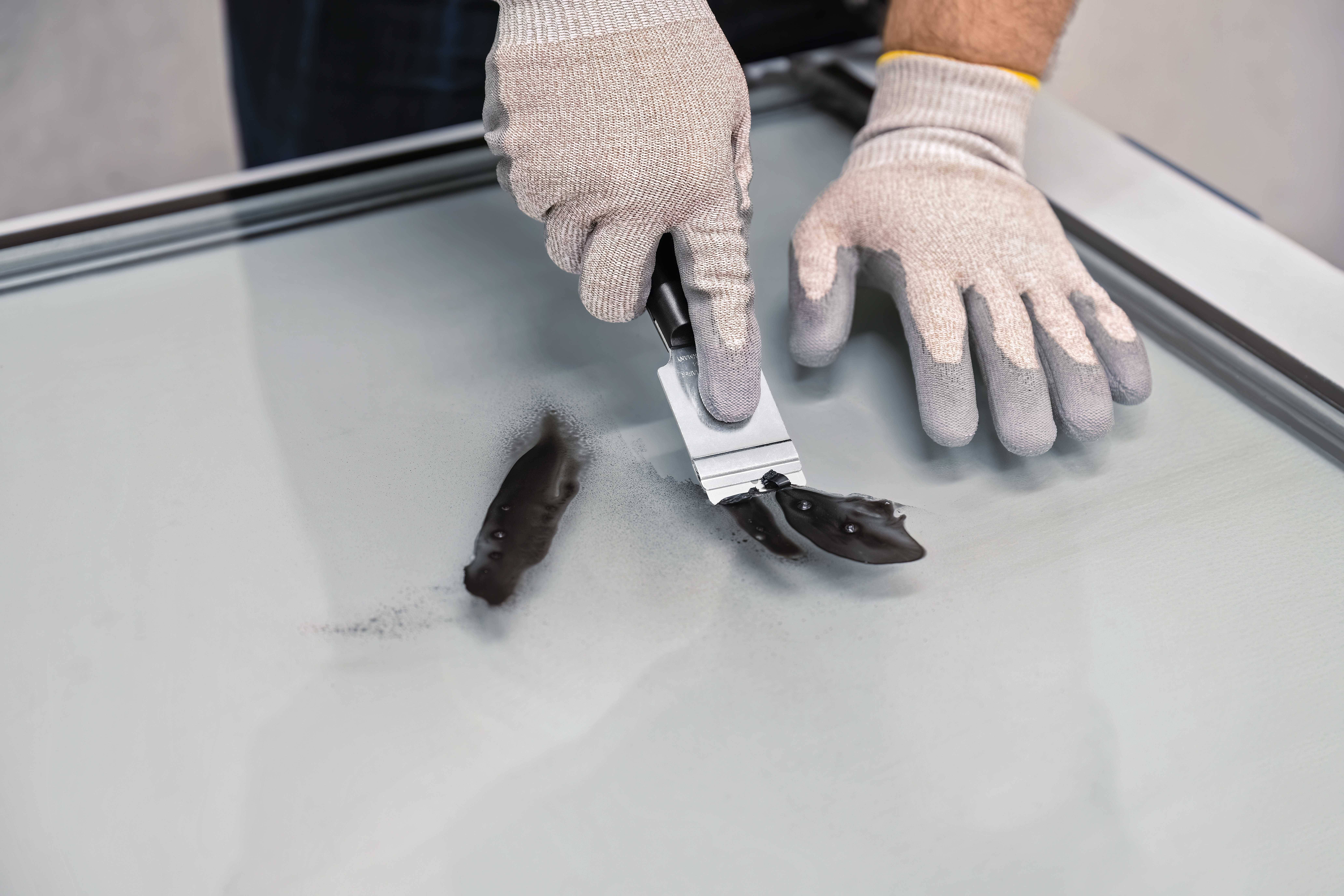 A person is wearing gloves and removing black marks from a glass surface using a scraper.
