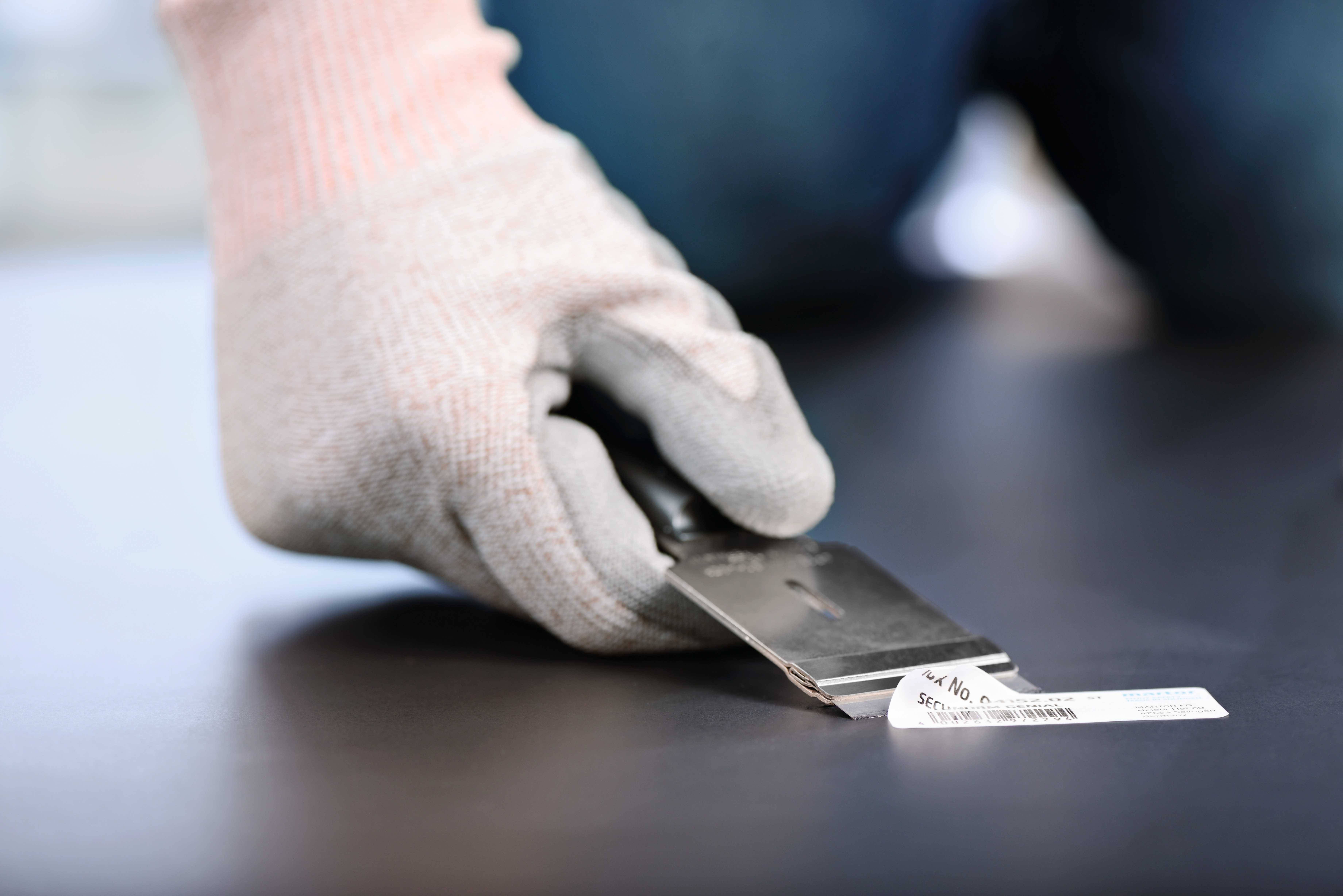 A person wearing grey gloves uses a blade to remove a label from a smooth surface.