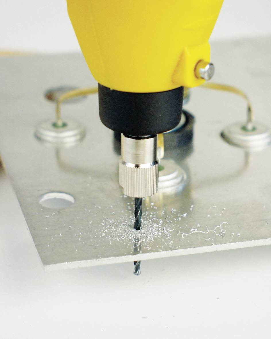 'A yellow drill is drilling a hole in a metal plate, with metal shavings around the hole; metal contacts are visible in the background.'