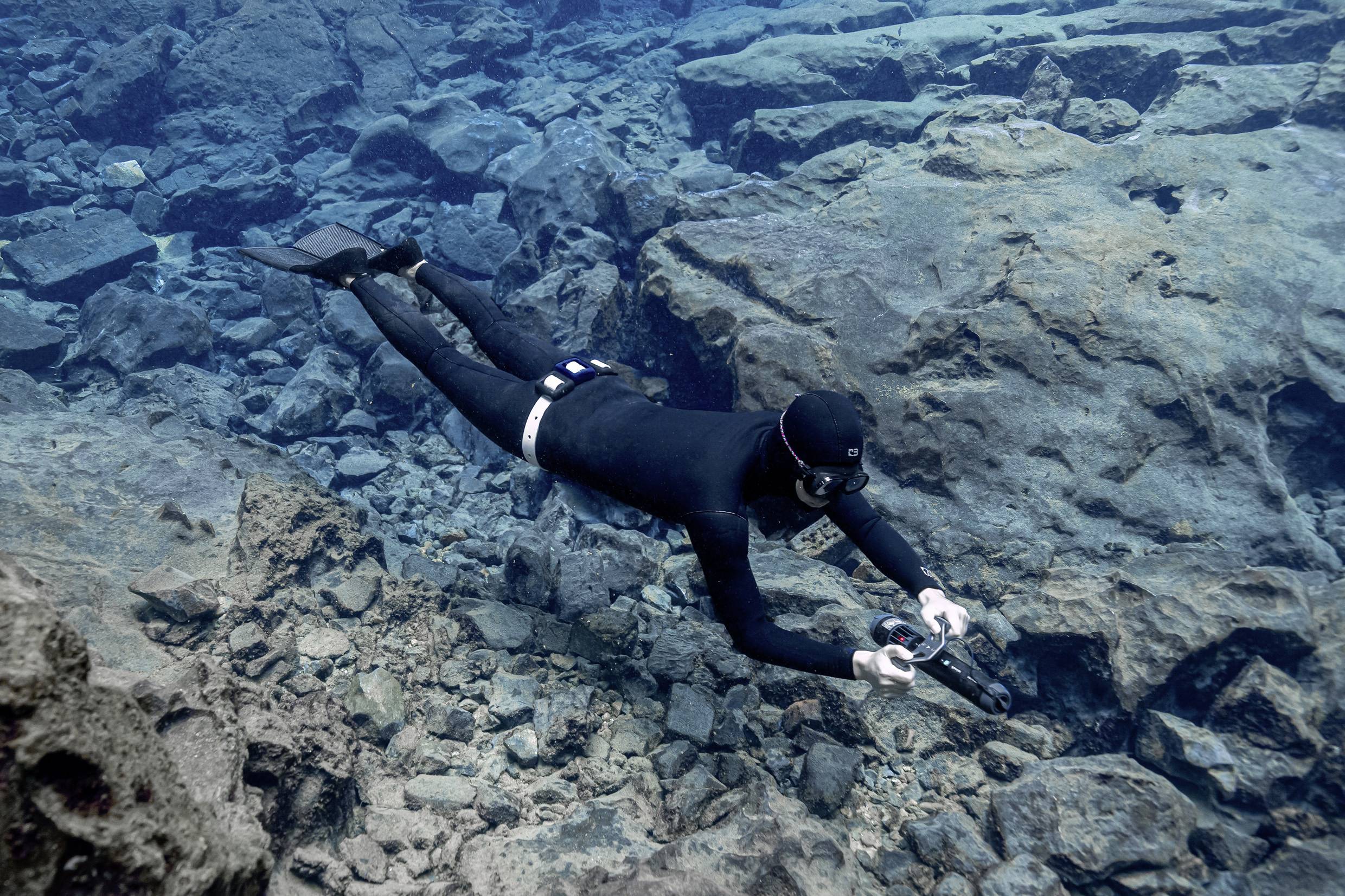 A diver in a black wetsuit swims between rocks underwater, holding a camera to capture underwater footage.
