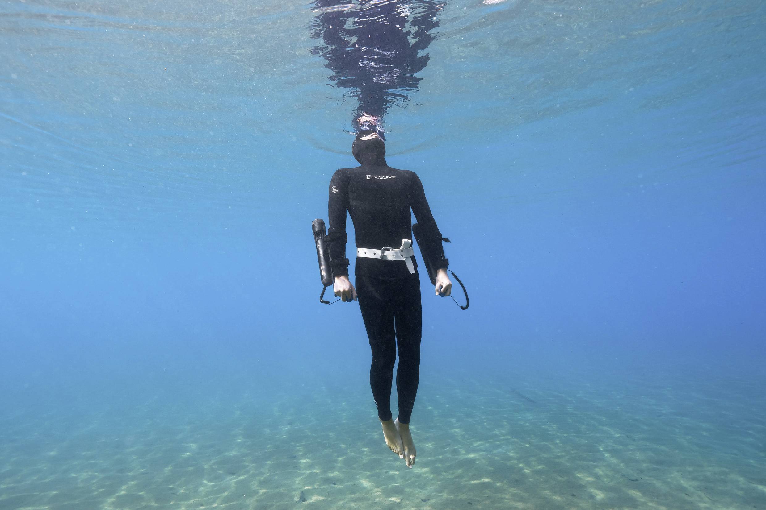 A person in a black wetsuit hovers underwater in a clear, blue sea, surrounded by tranquil water.