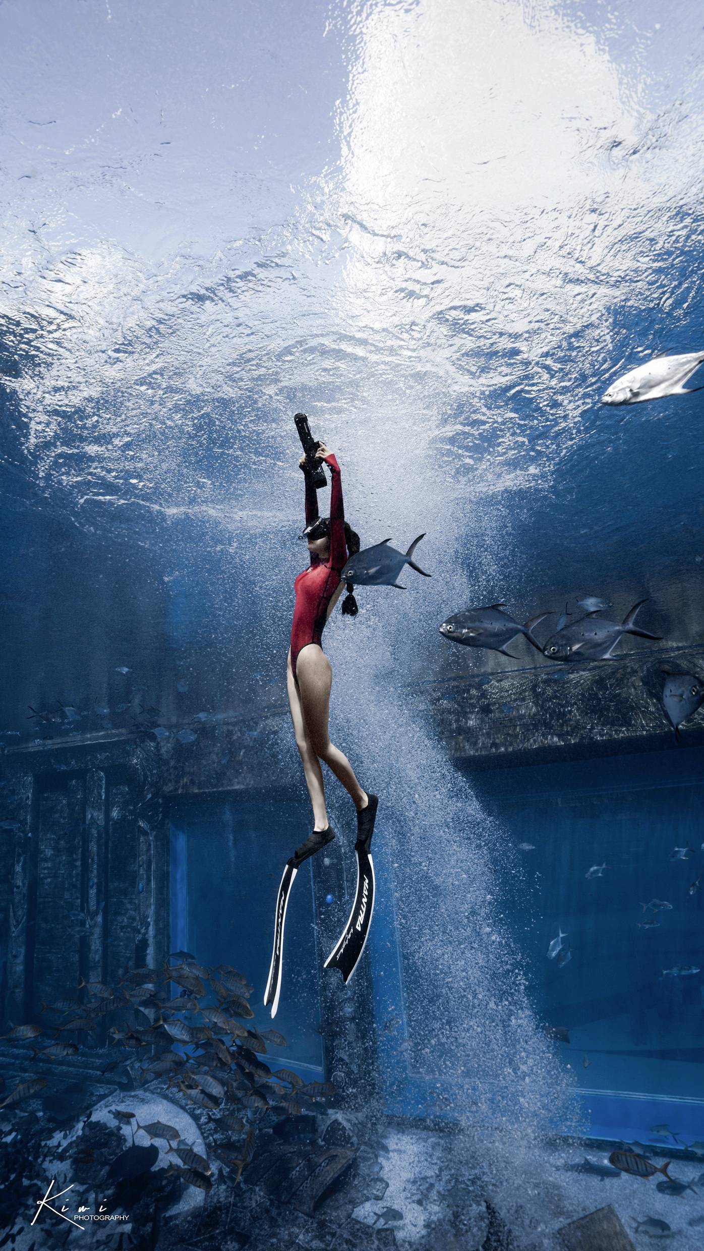 A person in a red diving suit is diving in a large aquarium with several fish, surrounded by clear water and ancient structures.
