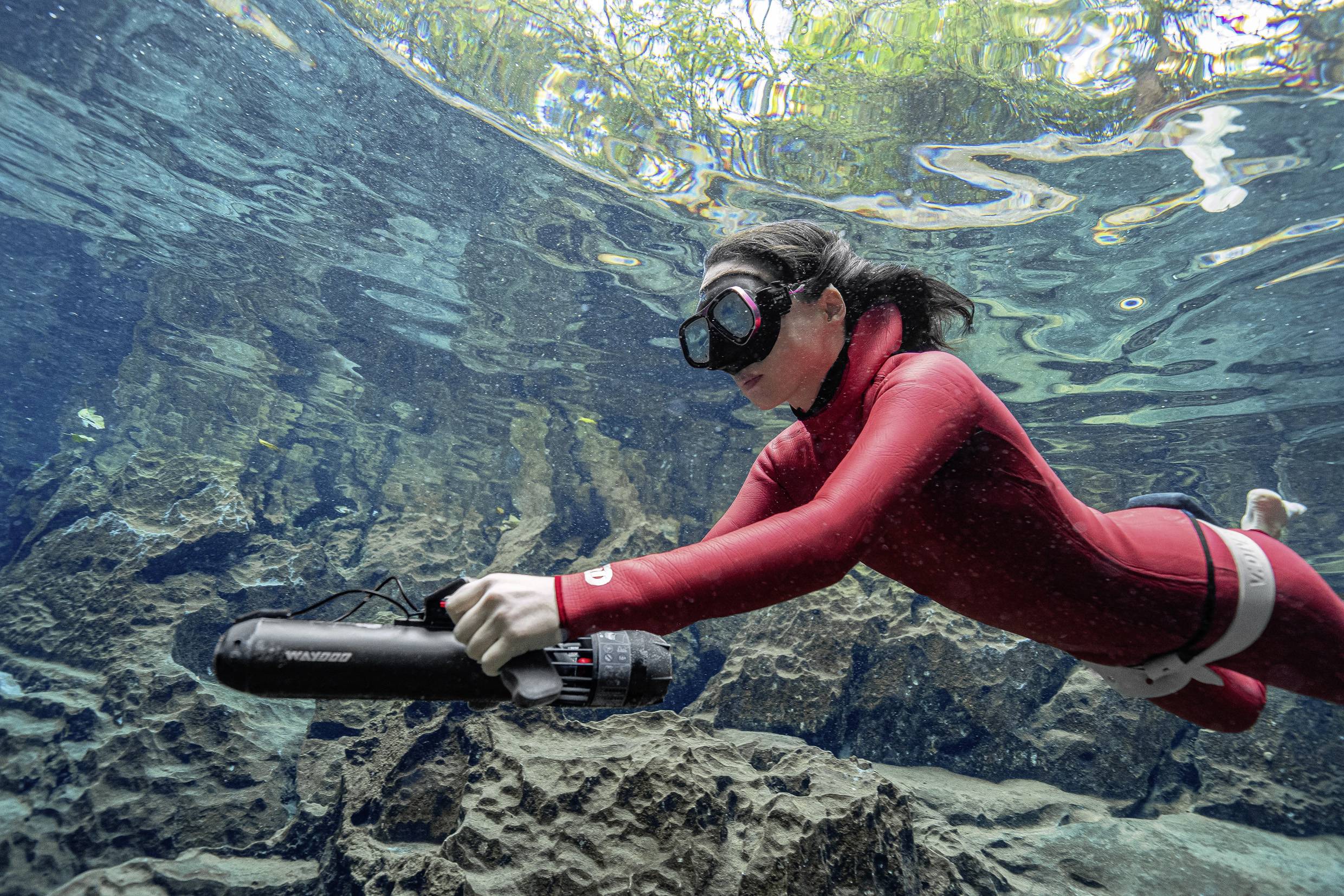 A person in a red diving suit is using an underwater scooter in clear water; rocky seabed and light reflections are visible.