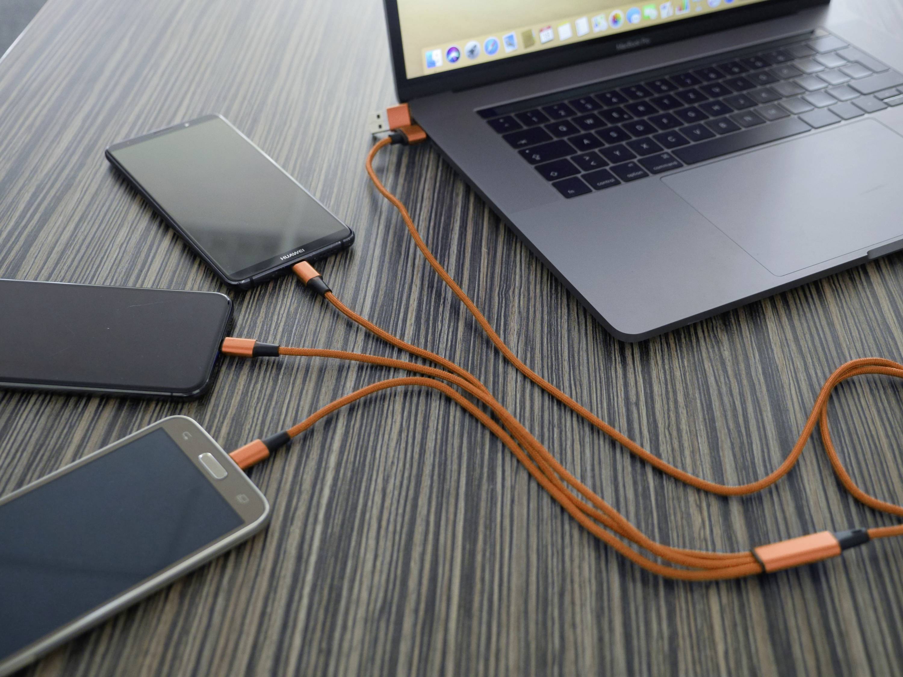 A laptop on a table is connected to three smartphones, all being charged via orange cables.