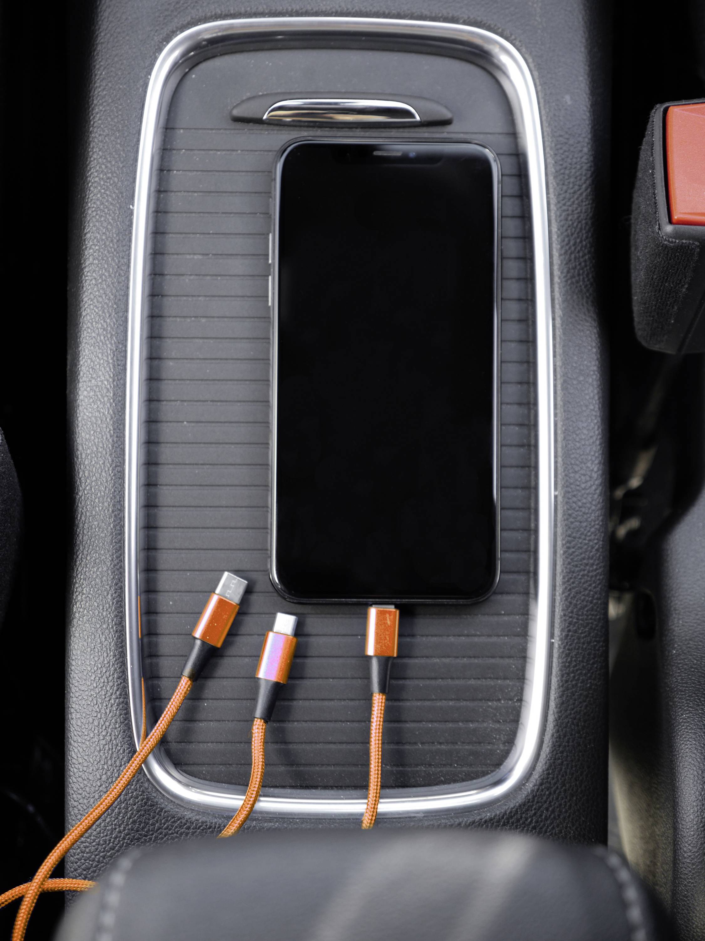 A smartphone is lying in a car storage compartment next to orange charging cables with various connectors.