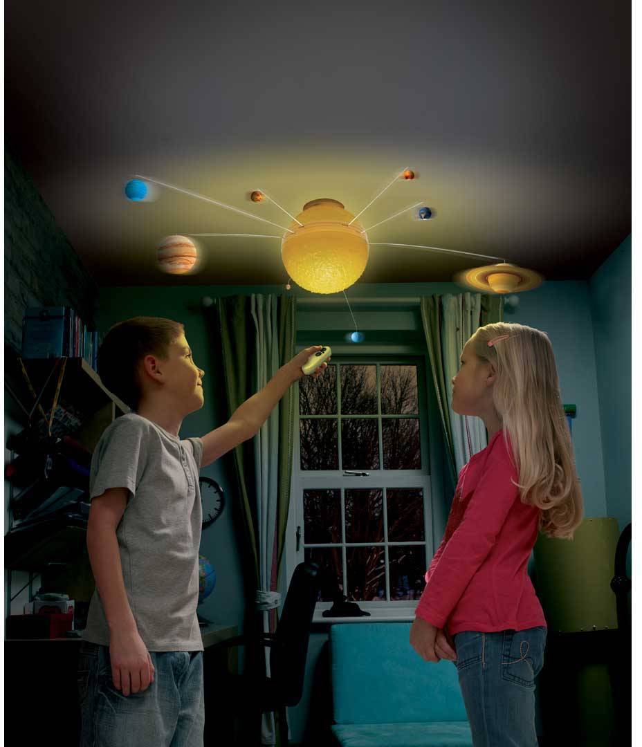 A boy and a girl are observing a glowing mobile solar system in a dark room. Planets are orbiting a sun lamp.