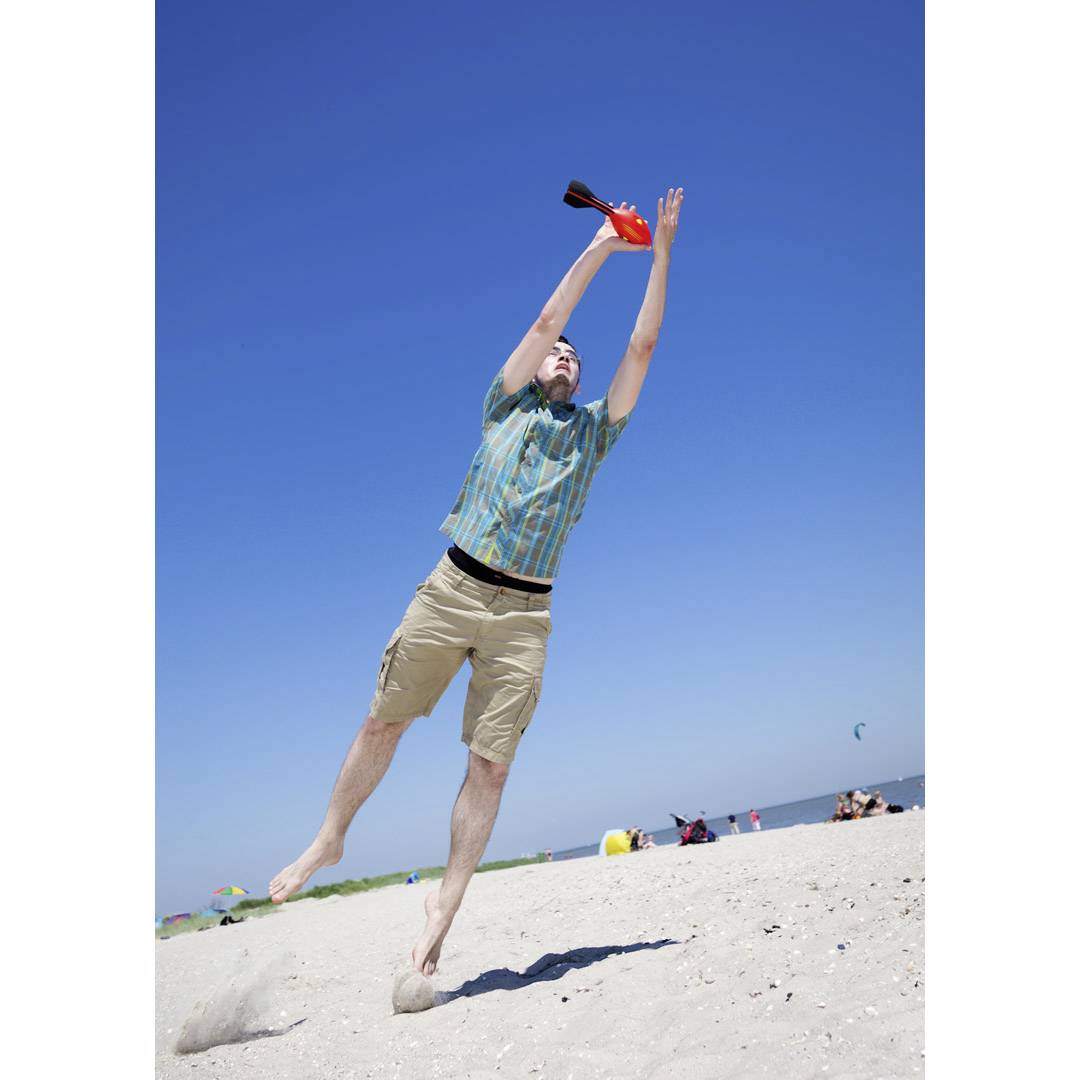 A man jumps on the beach and catches an orange object. In the background, the blue sky and sea can be seen.