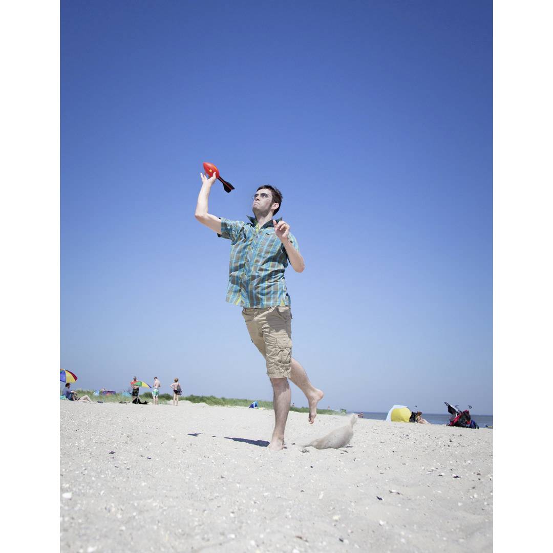 A man throws a frisbee on a sandy beach. The sky is clear and blue, with several people in the background.
