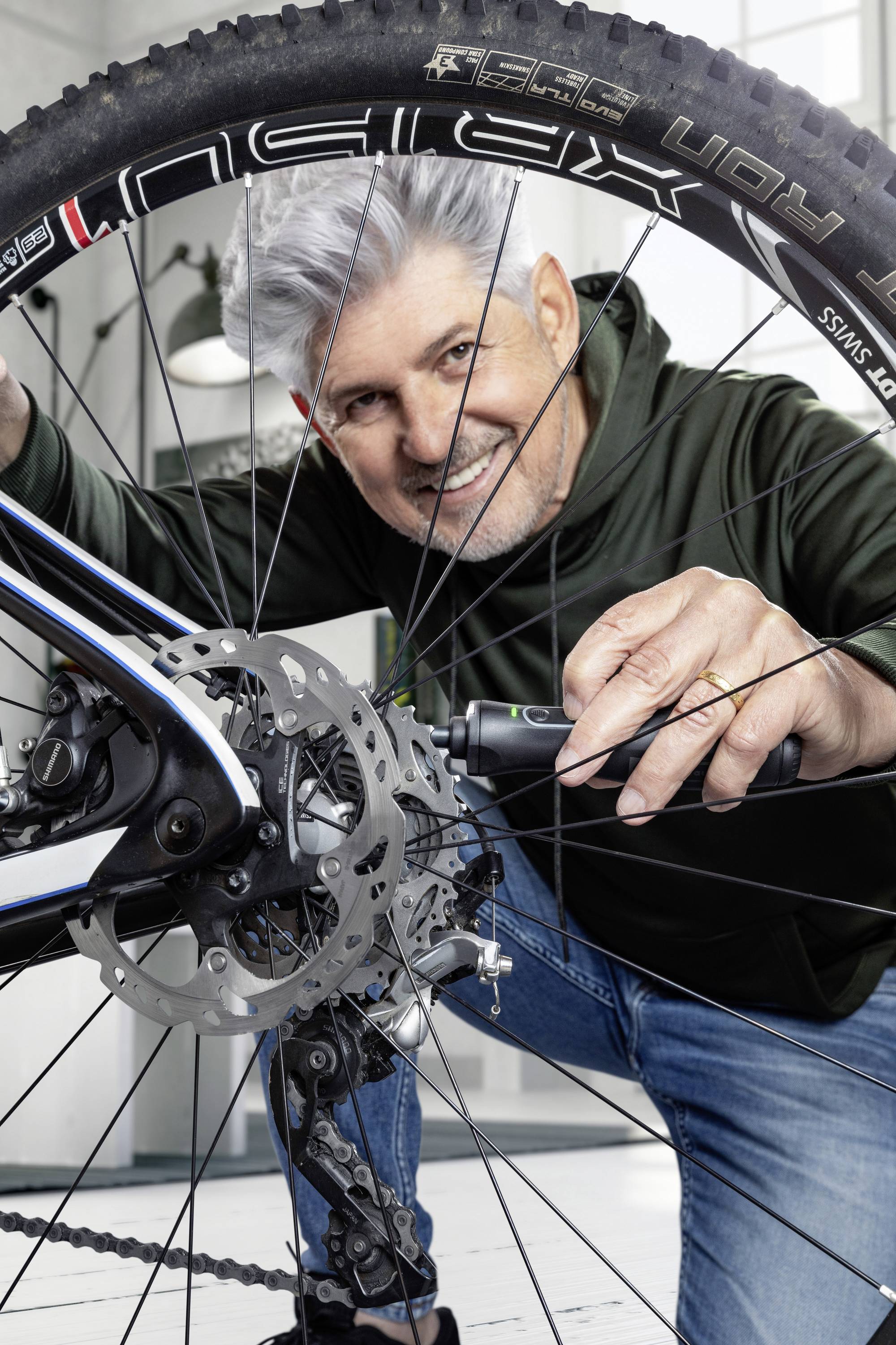 A man is repairing the rear wheel of a bicycle with a tool in his hand, smiling, and wearing a dark green hoodie.