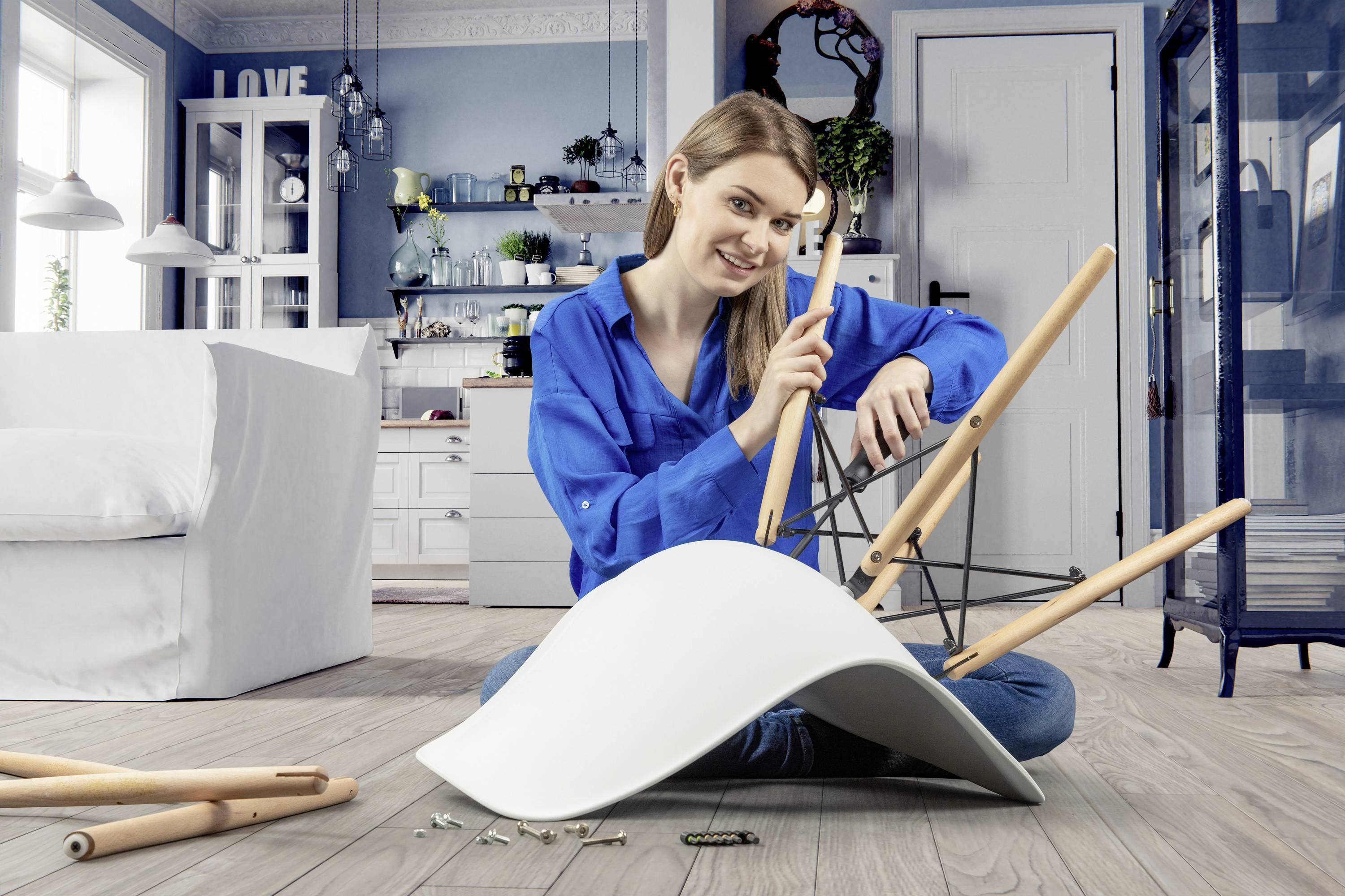 A woman in a blue shirt is sitting on the floor and assembling a white chair in a modernly furnished living room.