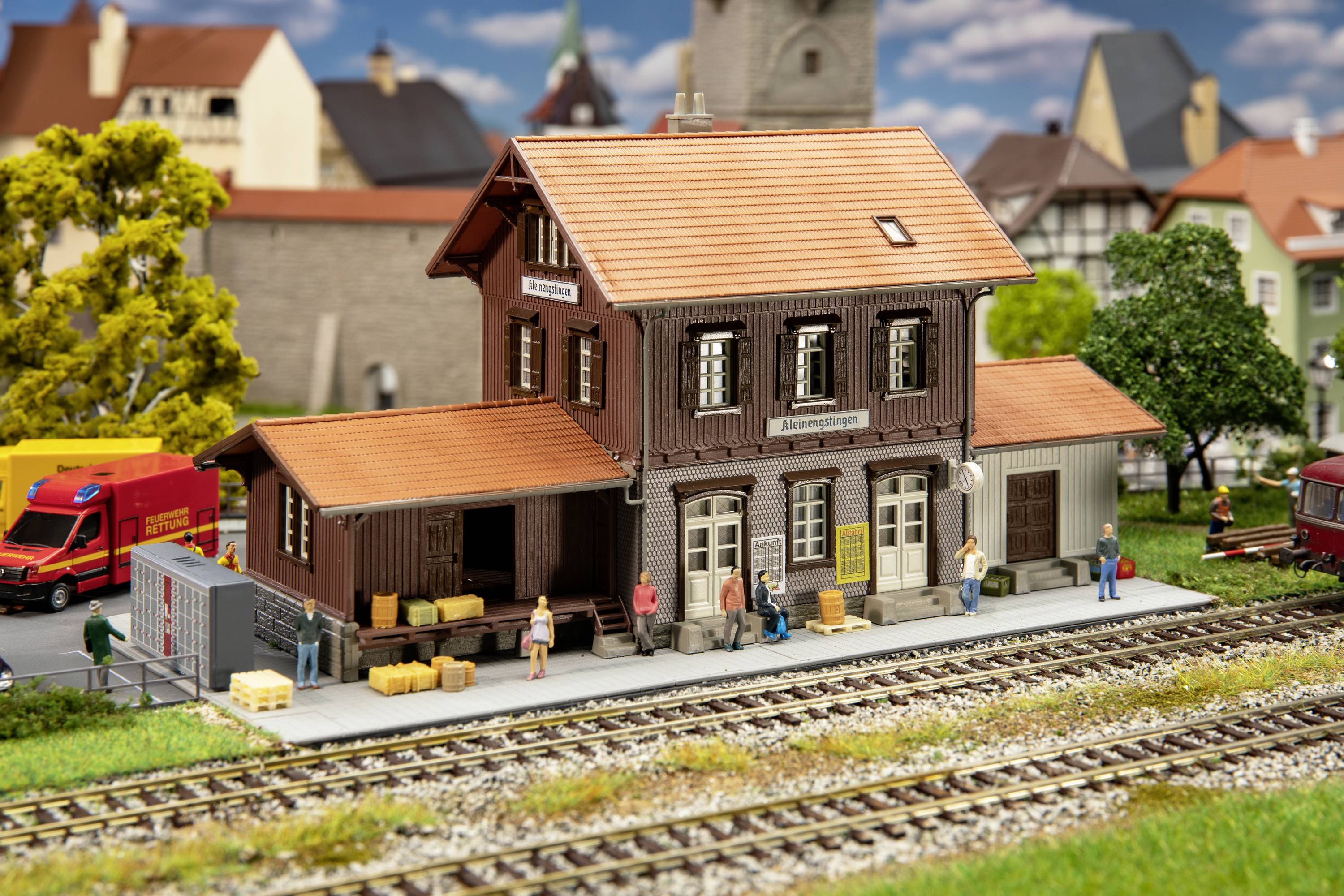 A model railway station in a rural setting with figurines, lorries, and railway tracks. The station has a two-storey building with wooden walls.