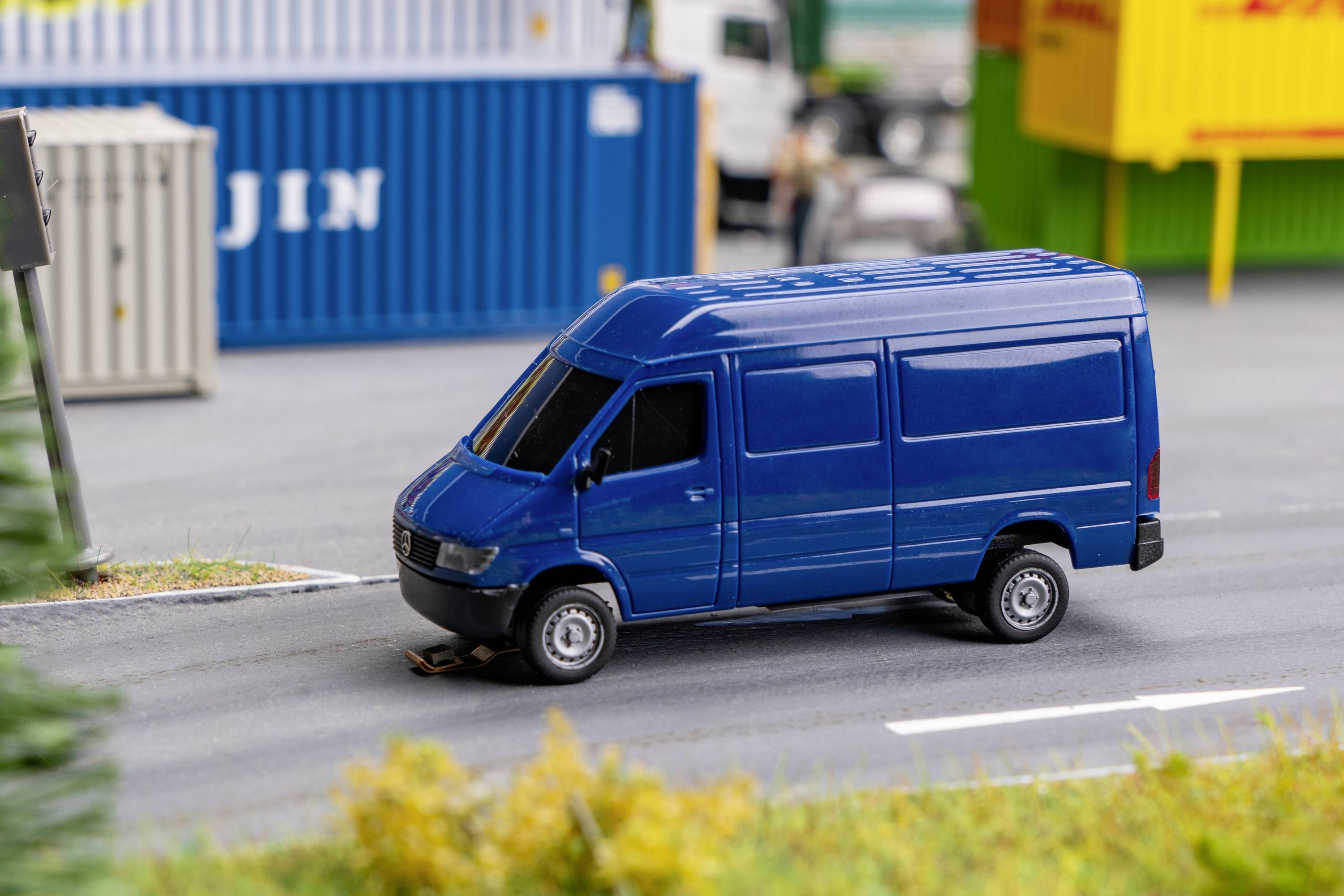A blue delivery van drives on a road, surrounded by freight containers. A scenario of an industrial area or harbour.