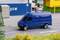 A blue delivery van drives on a road, surrounded by freight containers. A scenario of an industrial area or harbour.