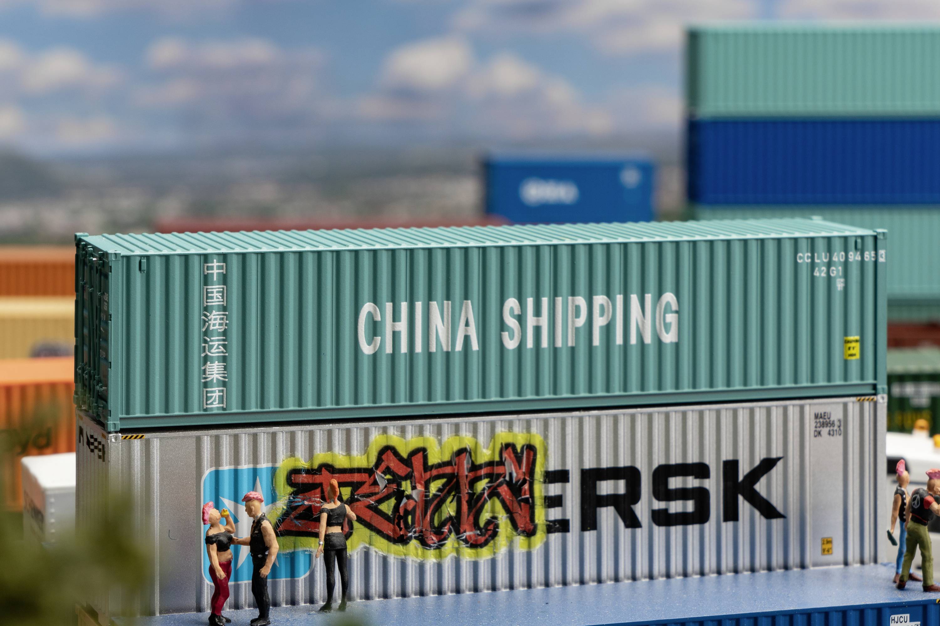Two stacked freight containers are visible, with 'China Shipping' on the top and 'Maersk' underneath, which is adorned with graffiti. People are surrounding the containers.