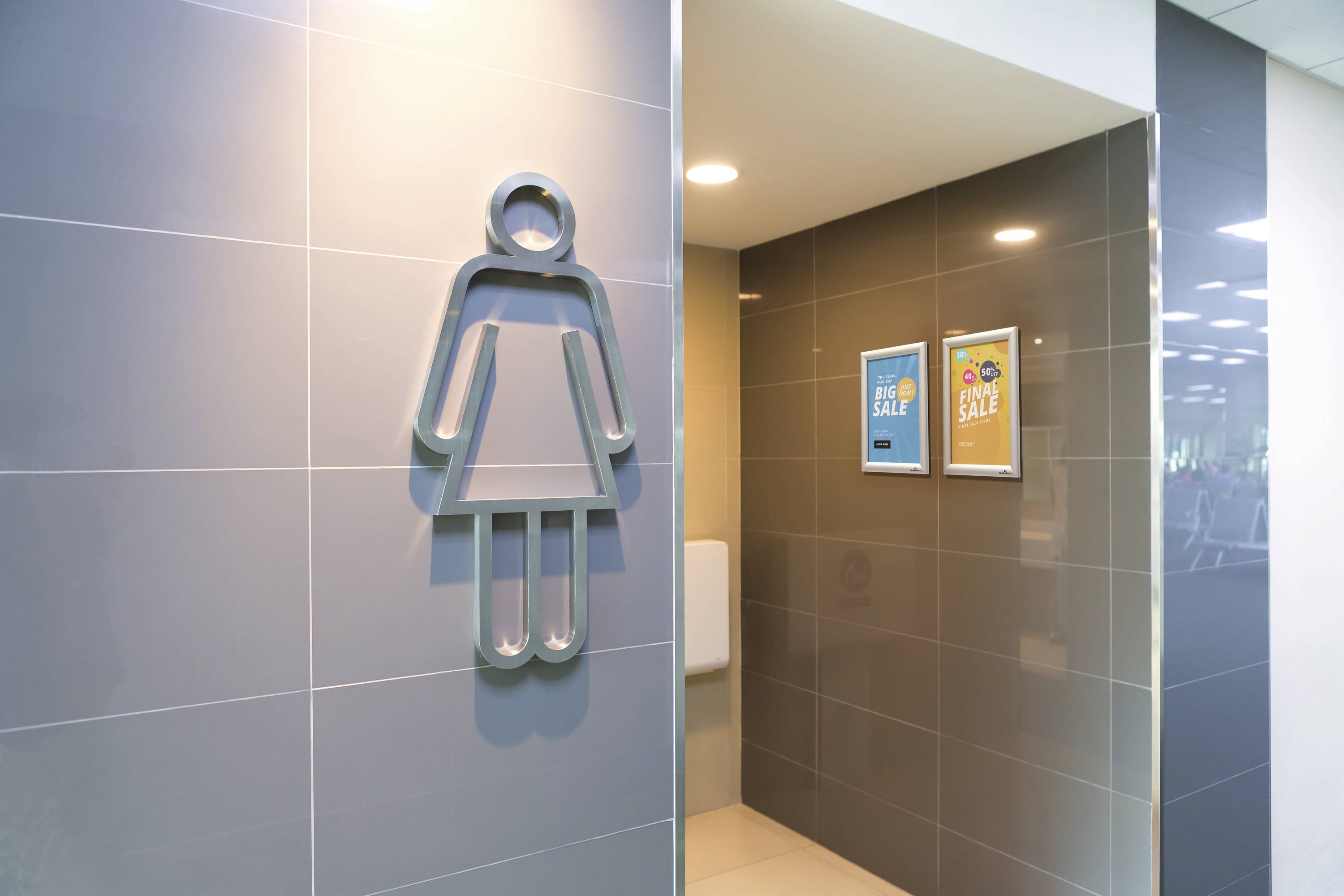 A woman indicates the symbol on the wall to the right, which denotes a female toilet area in a modern building.