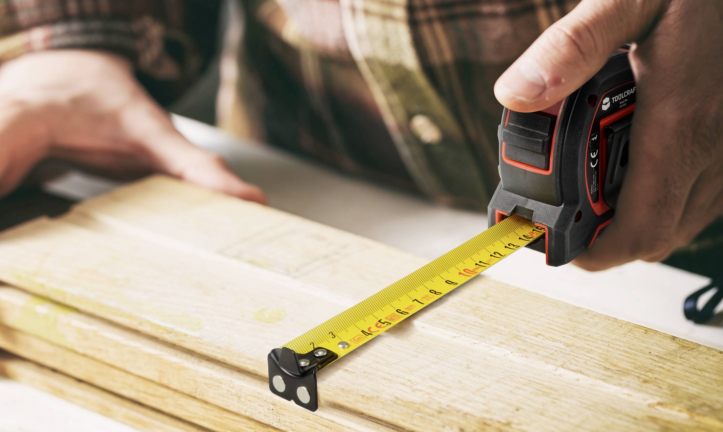 A person is measuring a wooden board with a tape measure. The scale shows approximately 26 centimetres.