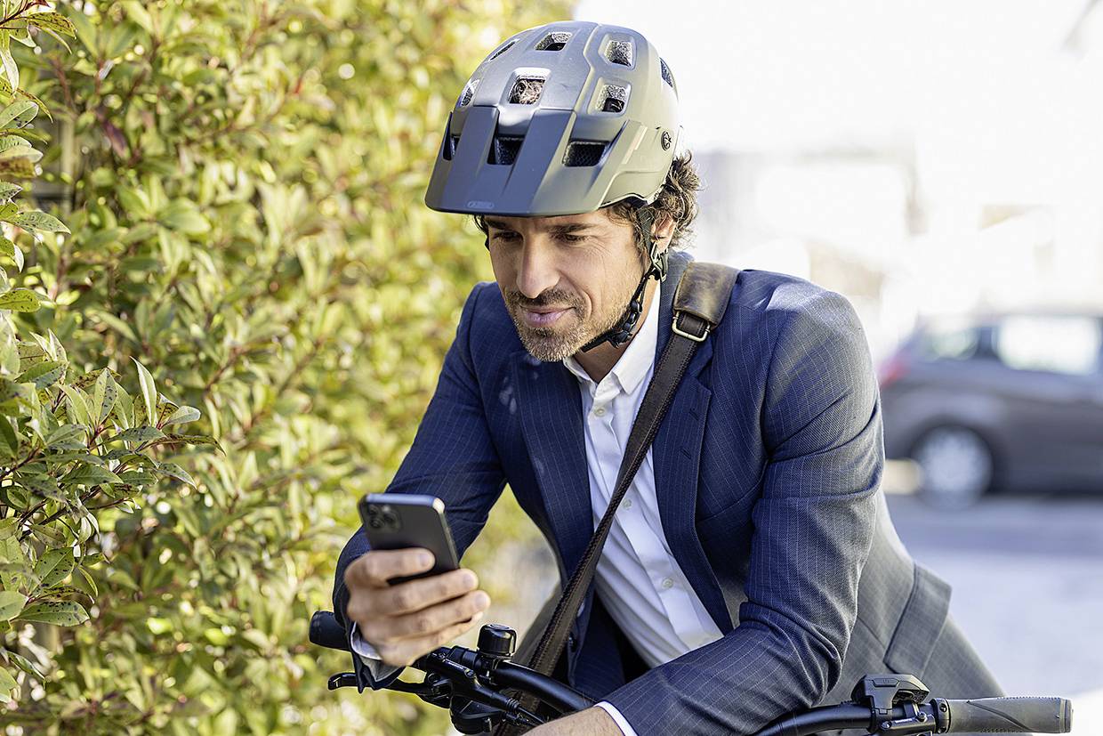 A man in a suit, wearing a helmet and holding a smartphone, sits on a bicycle beside a hedge on a road.