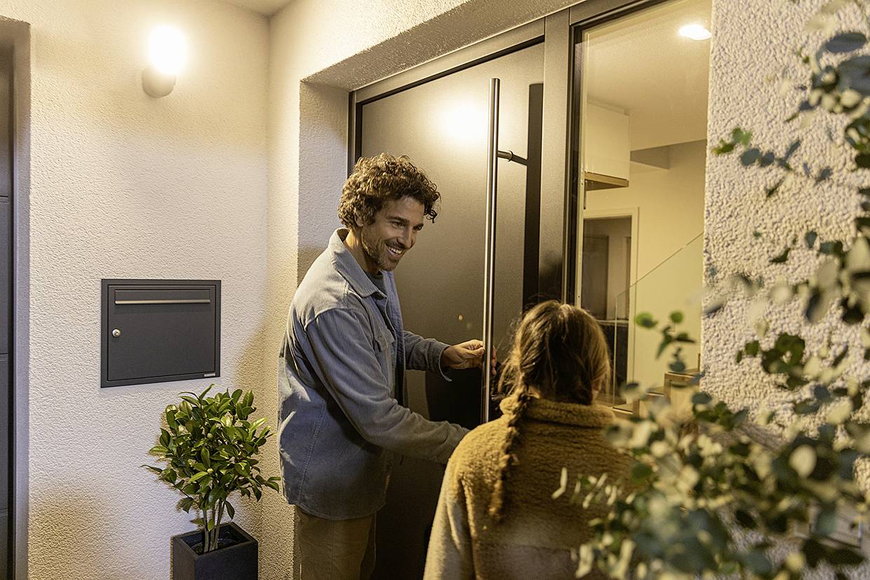 A man smiles as he opens the front door for a person with long hair. They are standing in front of an illuminated entrance with plants beside it.