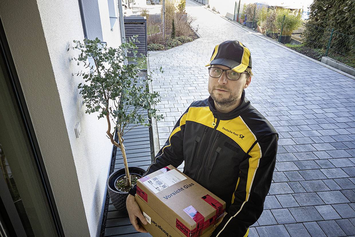 A delivery driver in a yellow and black uniform is holding a package in front of a front door with a modern driveway and a small potted tree.