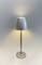 A modern, silver table lamp with a plain, cylindrical shade, emitting soft light towards the ceiling and floor.