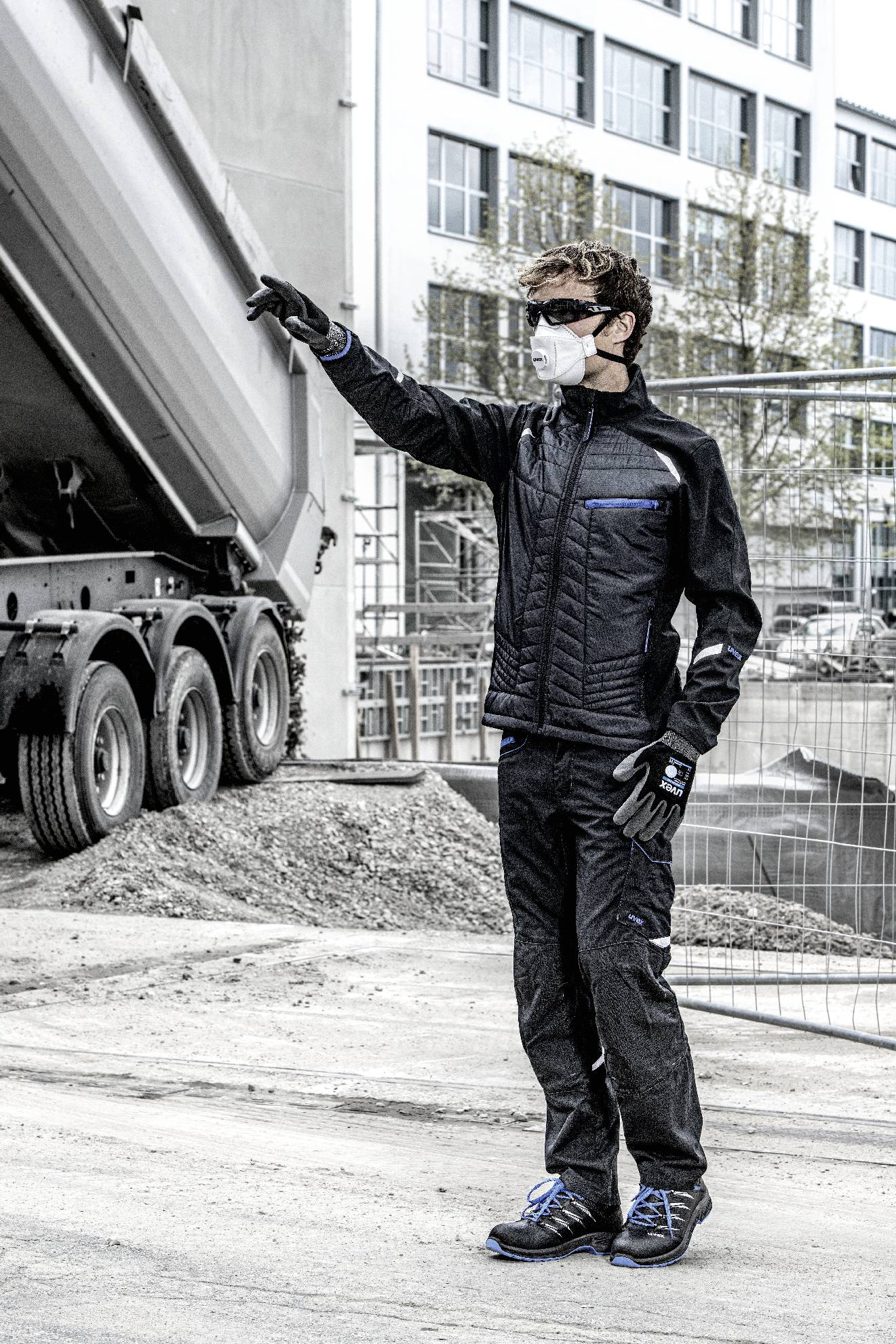 A person in workwear and a mask is pointing towards a construction site. A lorry is standing in the background. Buildings and construction fences are visible.
