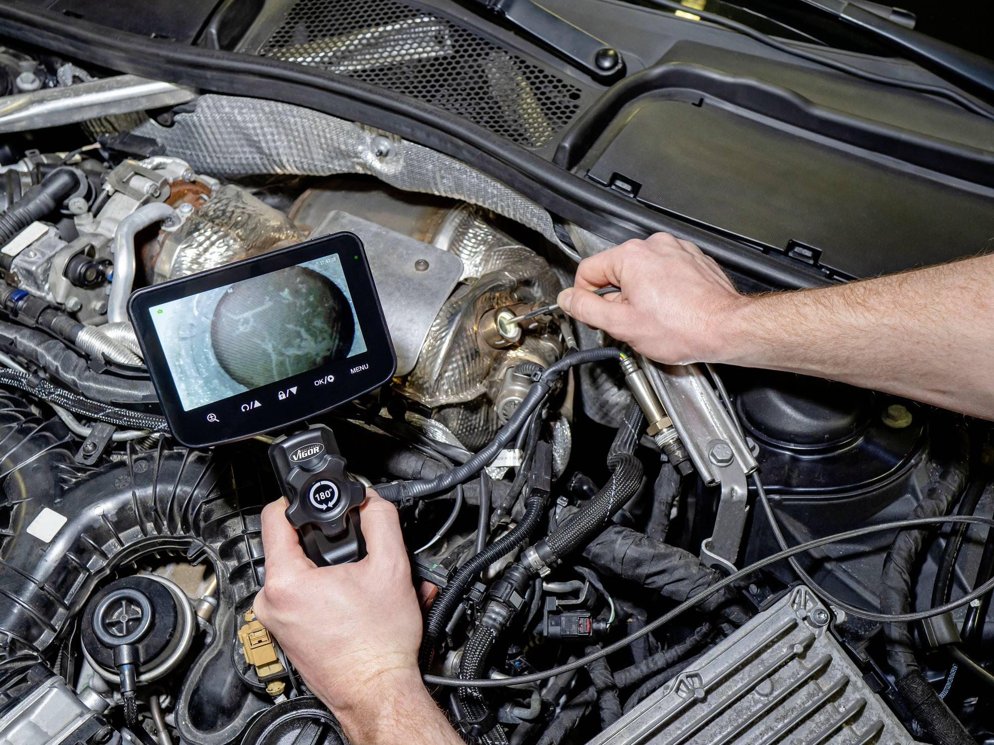 A person using a diagnostic camera to inspect an engine compartment, focusing on internal components. The camera display shows detailed imagery.