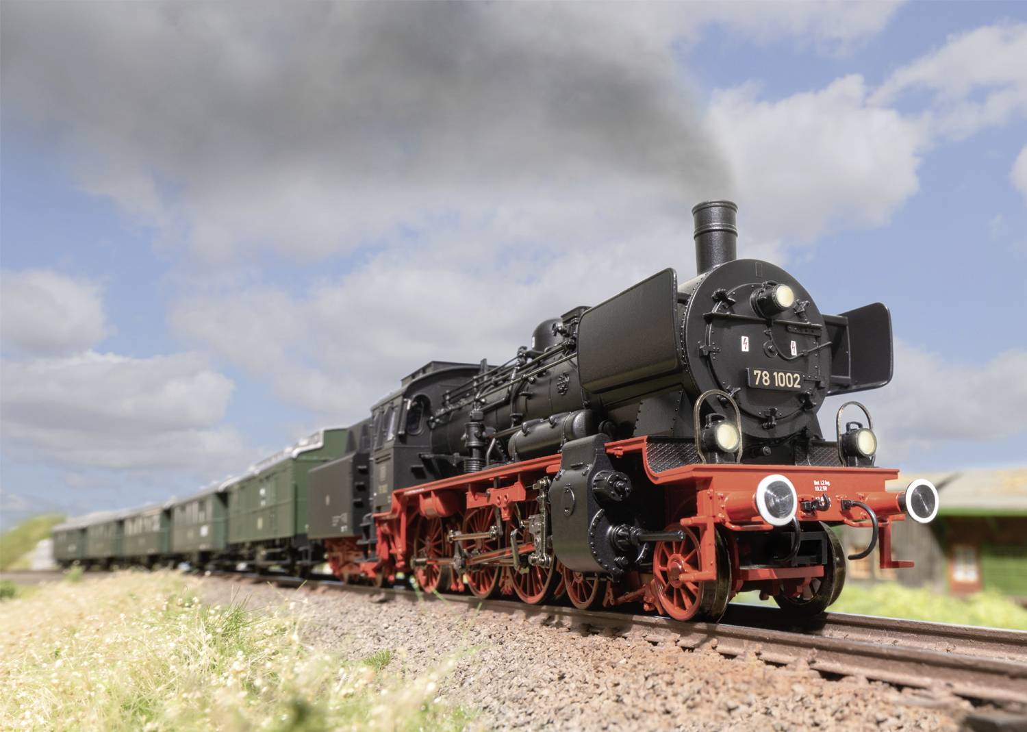 A black steam locomotive with a red chassis travels along a railway line through a rural landscape under a blue sky with clouds.