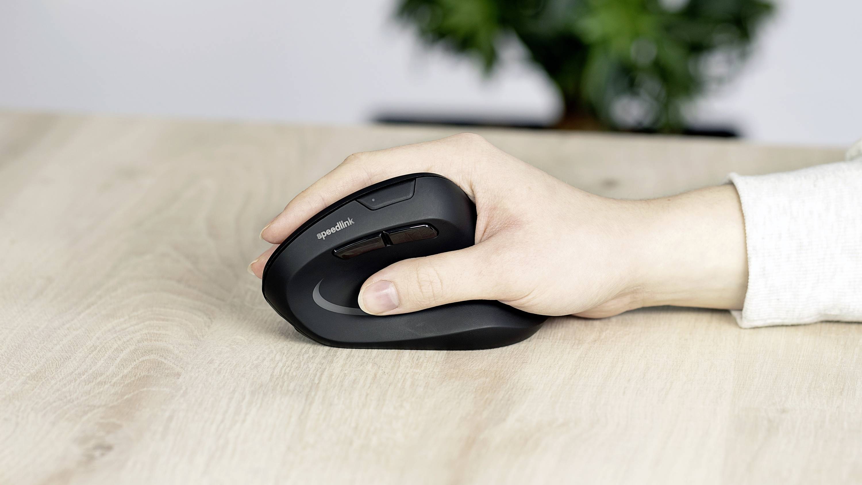 A hand is holding an ergonomic mouse on a wooden table. A plant is blurrily visible in the background.