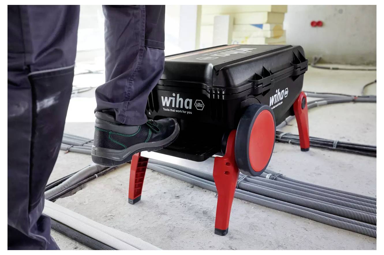 'Person stepping on a Wiha toolbox with integrated wheels and handles in a construction setting, surrounded by cables and pipes.'
