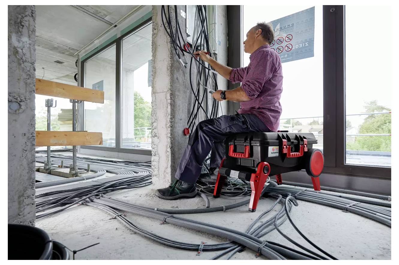 A person in a construction setting manages multiple cables attached to a wall. They are seated on a portable tool case, surrounded by tools.