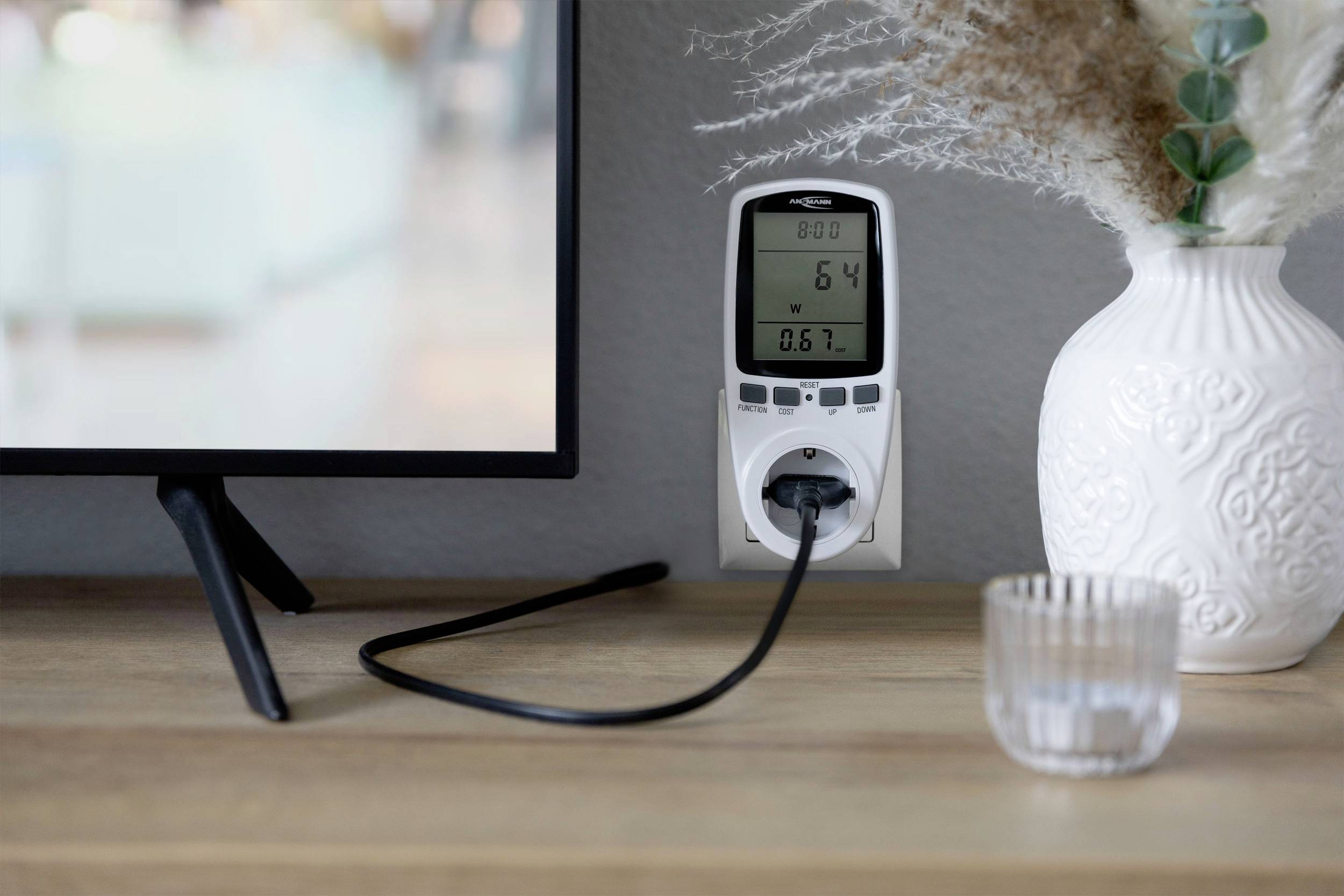 An electricity meter displays consumption data at a socket next to a television. Beside it stands a decorative vase.