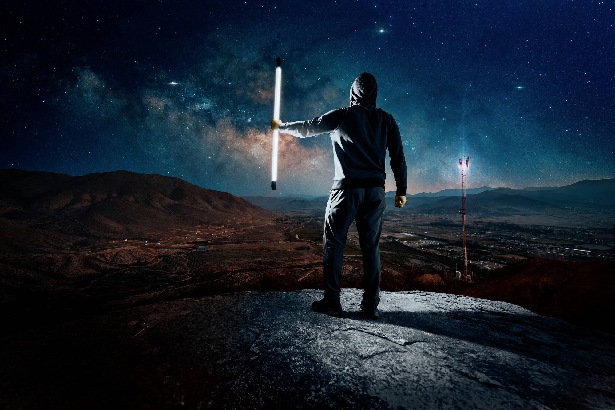 A person in a hoodie stands on a rock, holding a glowing tube at night, gazing out over a vast landscape beneath a starry sky.