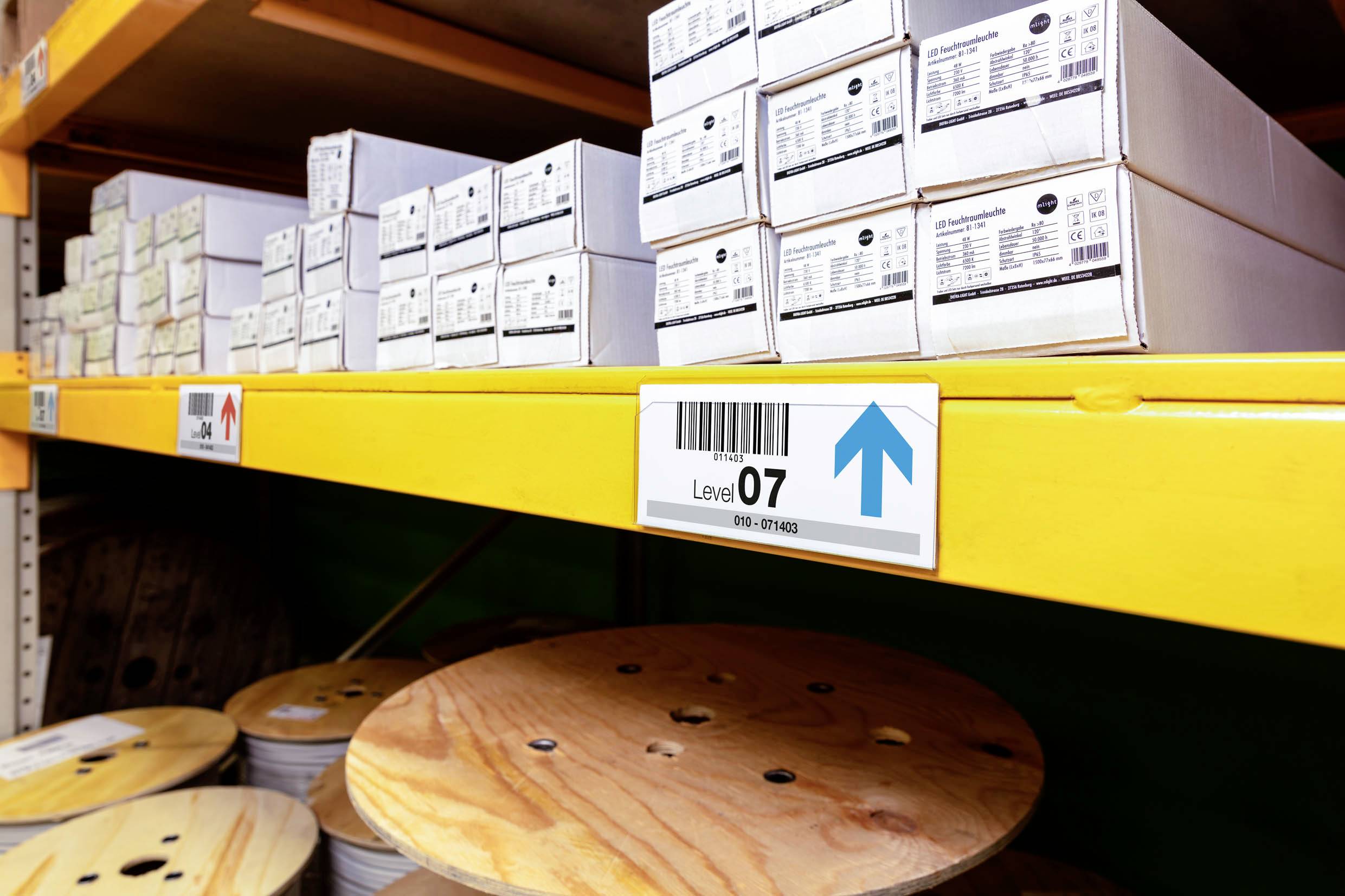 Shelving with white boxes, labelled 'Level 07', with a blue upward arrow. Cable reels on the floor. Warehouse environment.