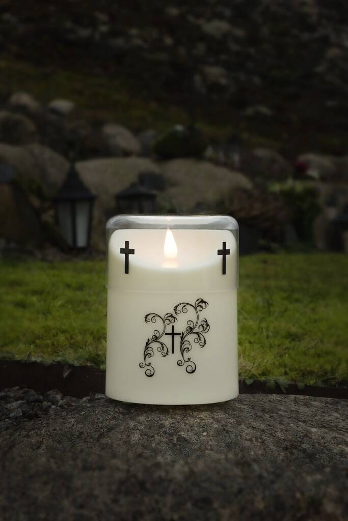 A burning candle with a cross and floral decorations sits on a stone surface outdoors, surrounded by blurred natural scenery.