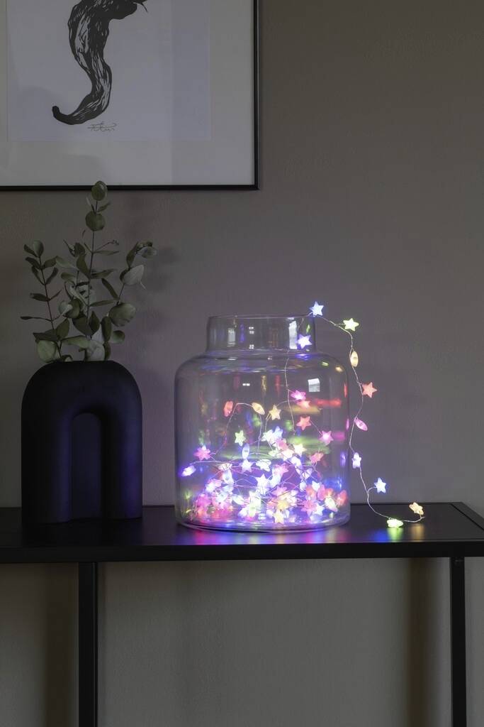 A glass vase on a table contains colourful glowing starry lights. Beside it stands a black vase with eucalyptus branches.
