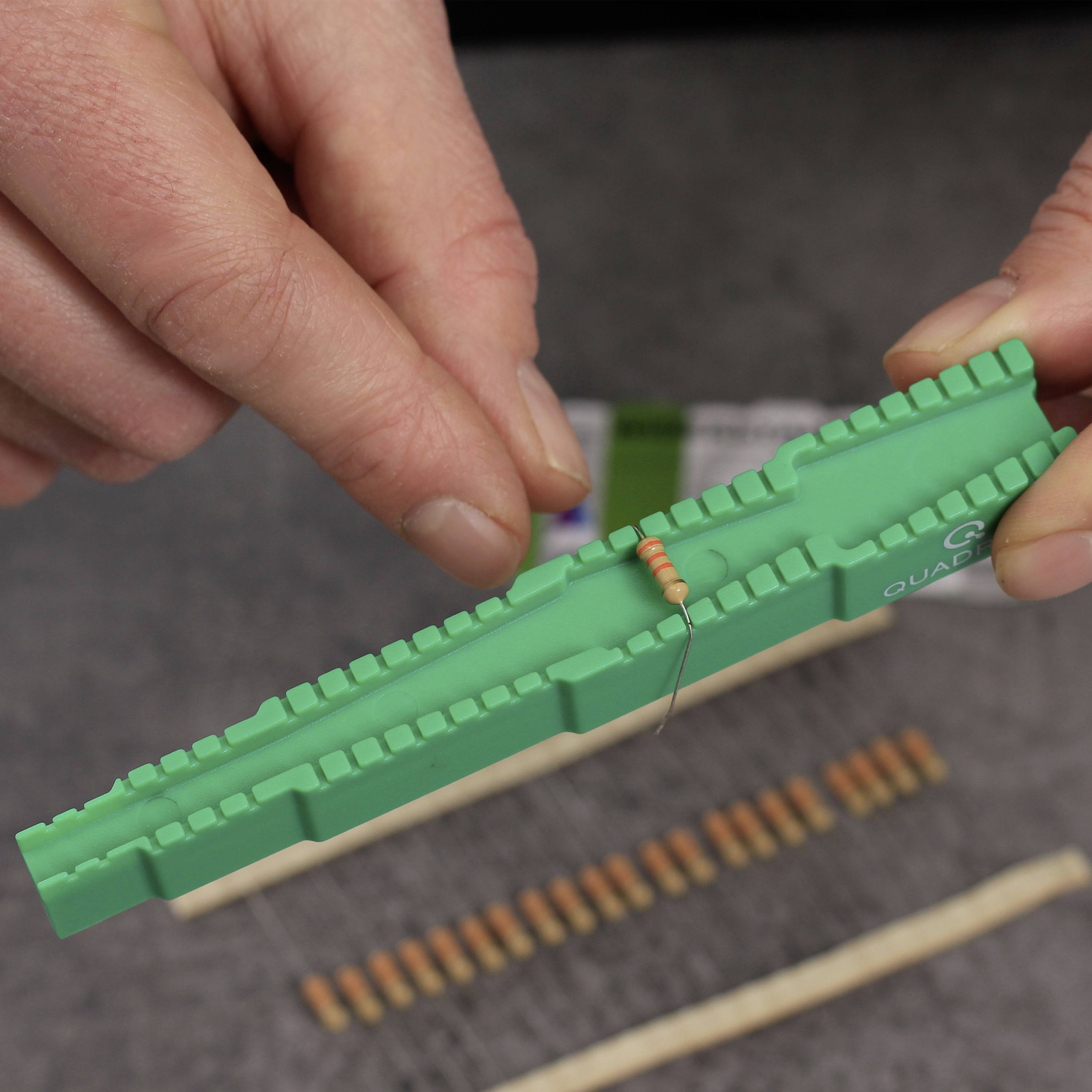A person is holding a green electronic component holder with a resistor passed through it. Further resistors are lying in the background.