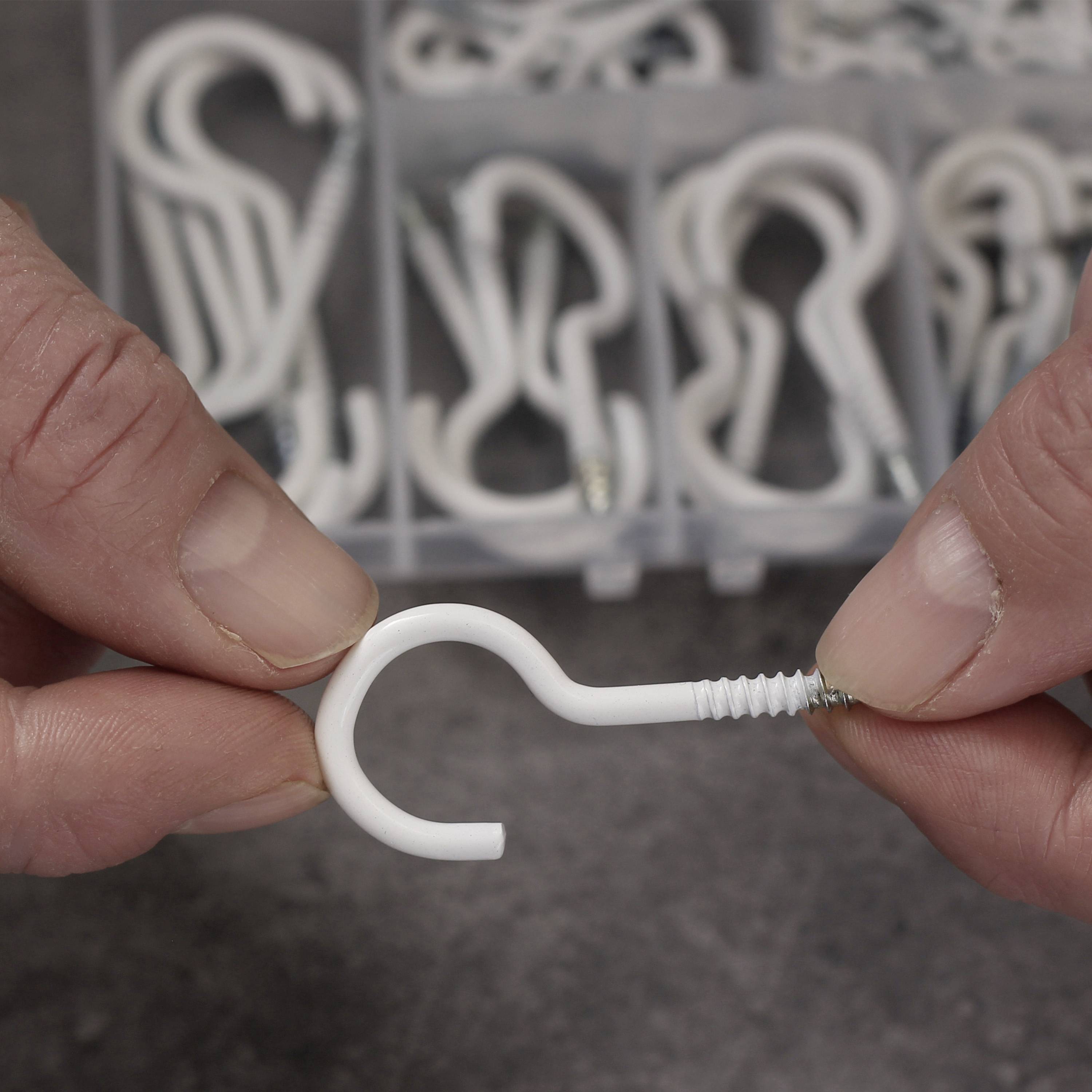 A hand is holding a white screw hook. In the background, many similar screw hooks can be seen in a storage box.