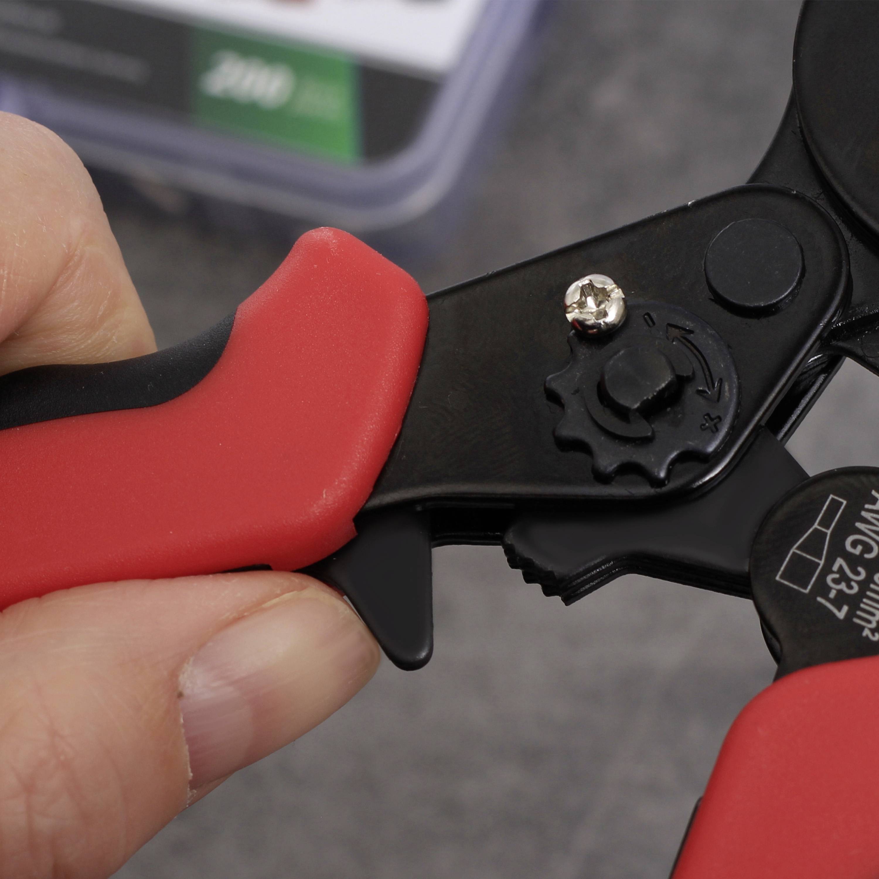 A hand is holding pliers with a red handle, turning a small black wheel on a wire. A toolbox is visible in the background.