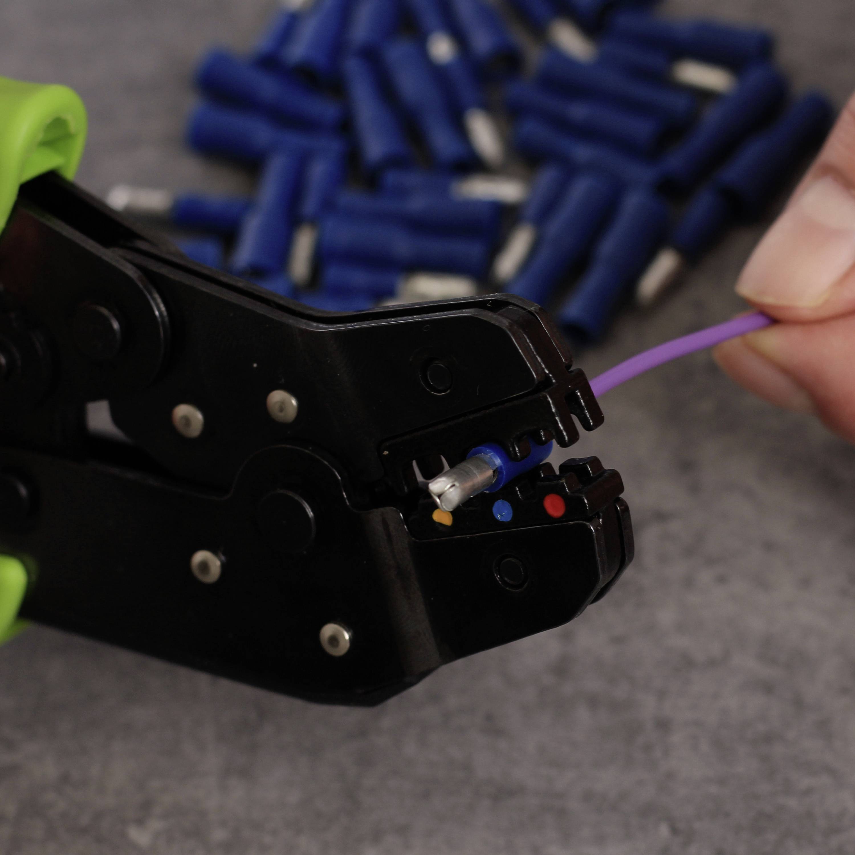 A crimping plier connects a purple cable with a blue cable lug. Blue cable lugs are lying in the background.