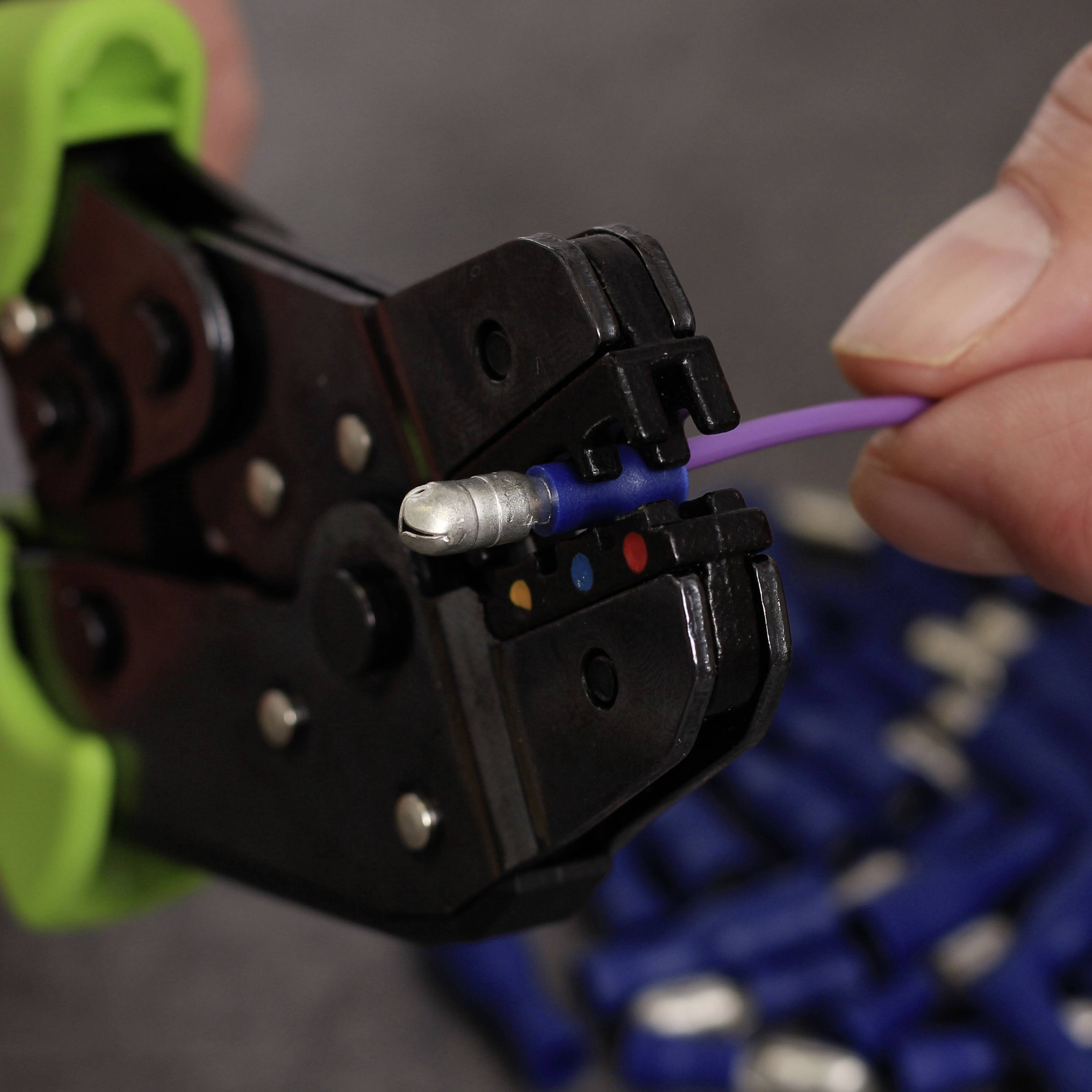 A hand holds a wire being inserted into a crimping tool. The tool has green handles and is positioned against a blue background.