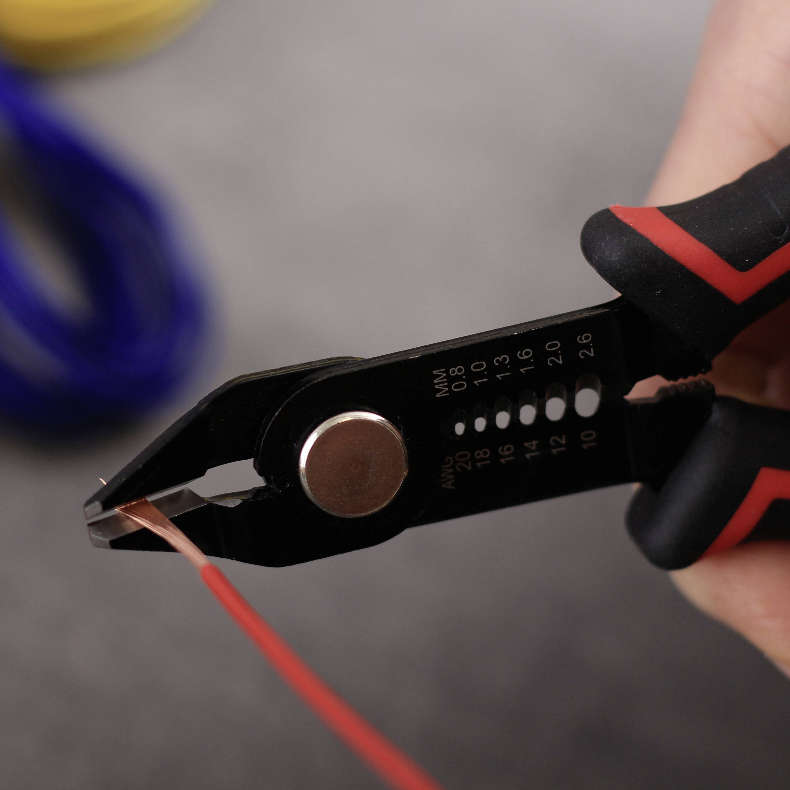 A hand holds wire strippers cutting an insulated wire. Blurred cables are visible in the background.