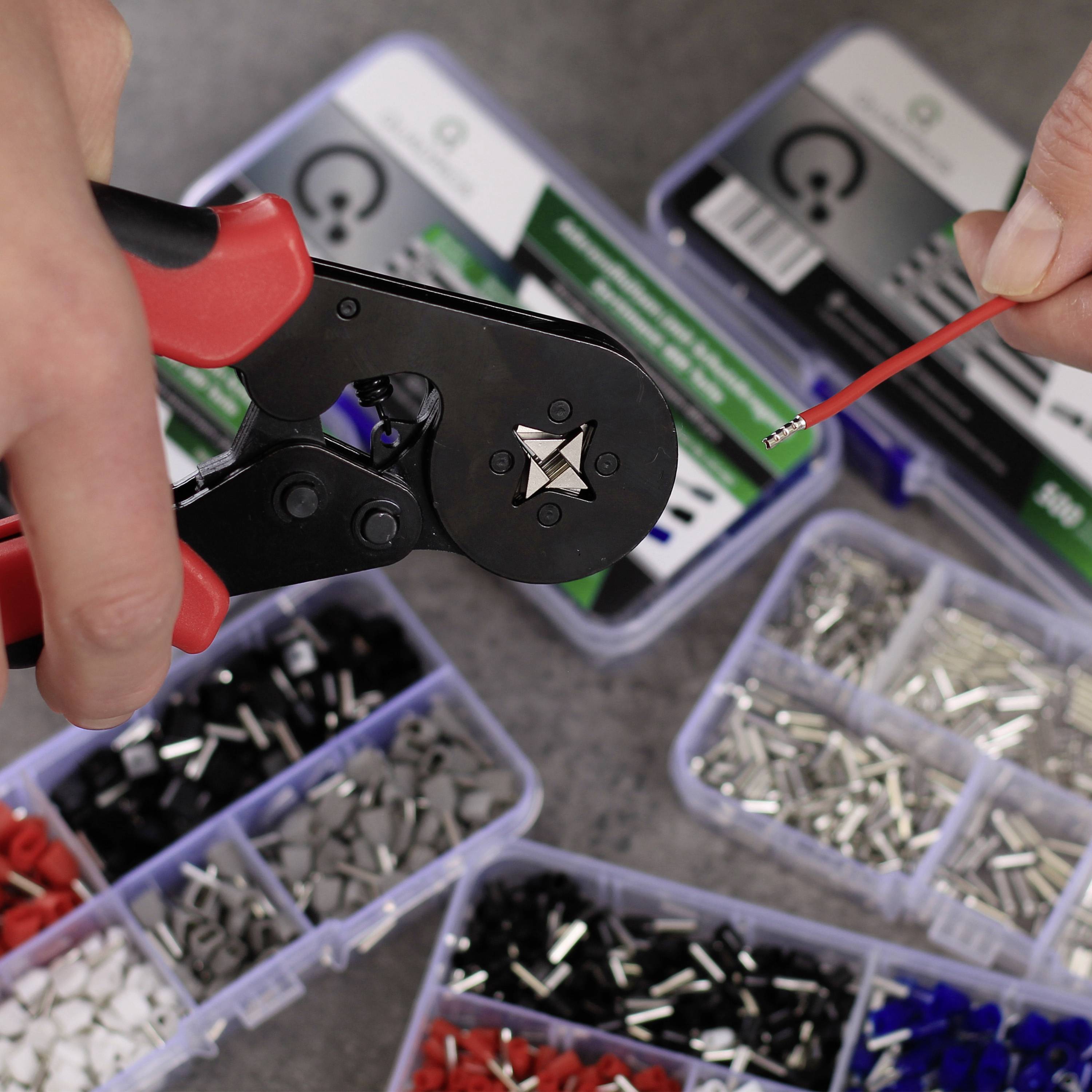 A hand holds a crimping plier working on a red wire. In the background, various plastic containers with cable clips are visible.
