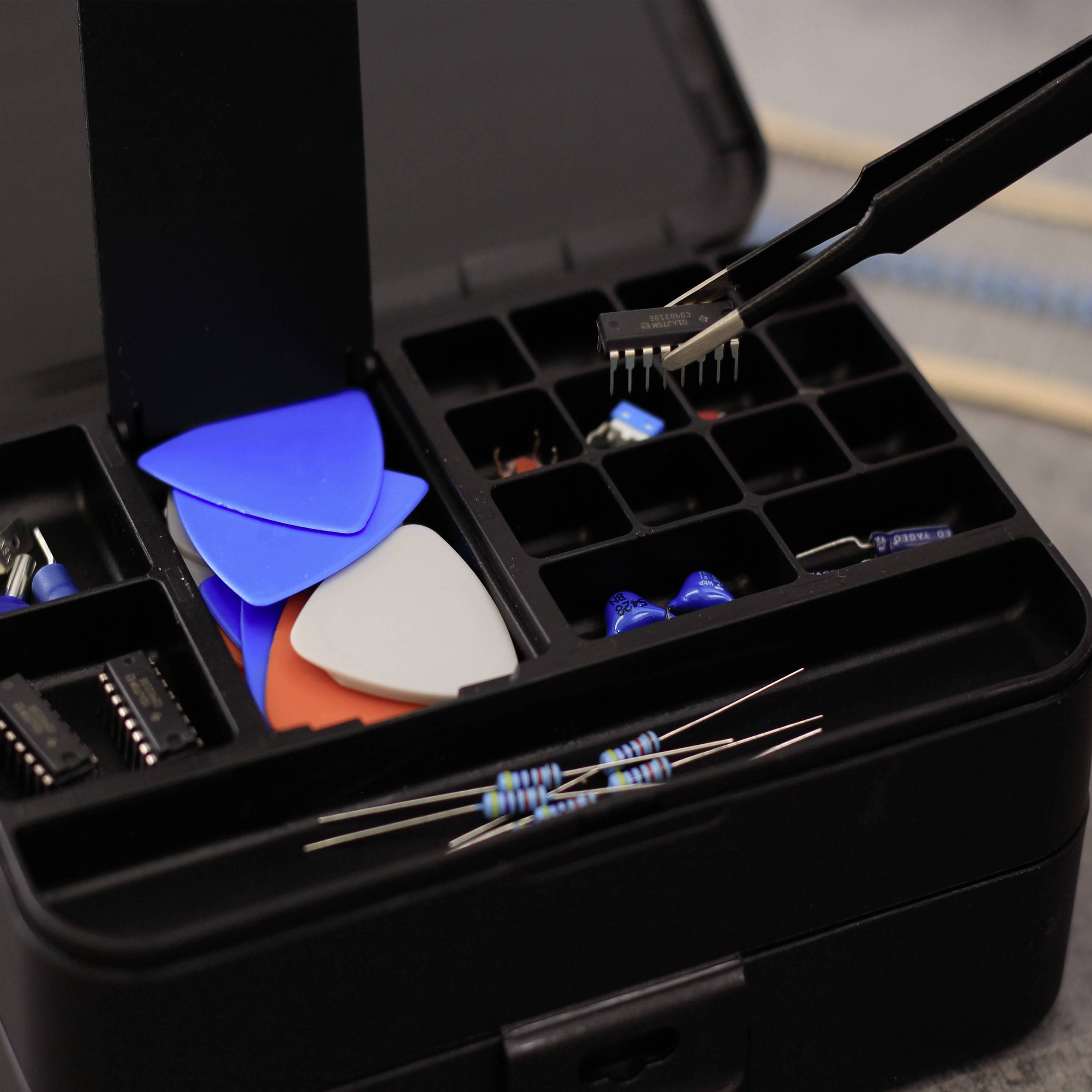 An open toolbox with electronic accessories: colourful plectrums, resistors and a pair of tweezers holding a small component.