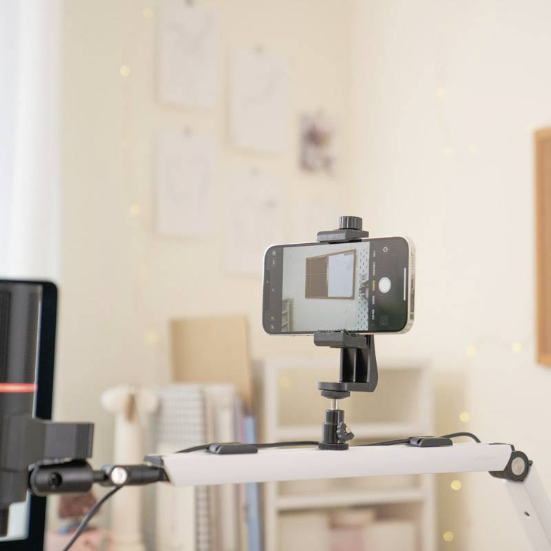 A smartphone is mounted on a tripod and shows the screen of a computer. Blurry shelves and pictures are visible in the background.
