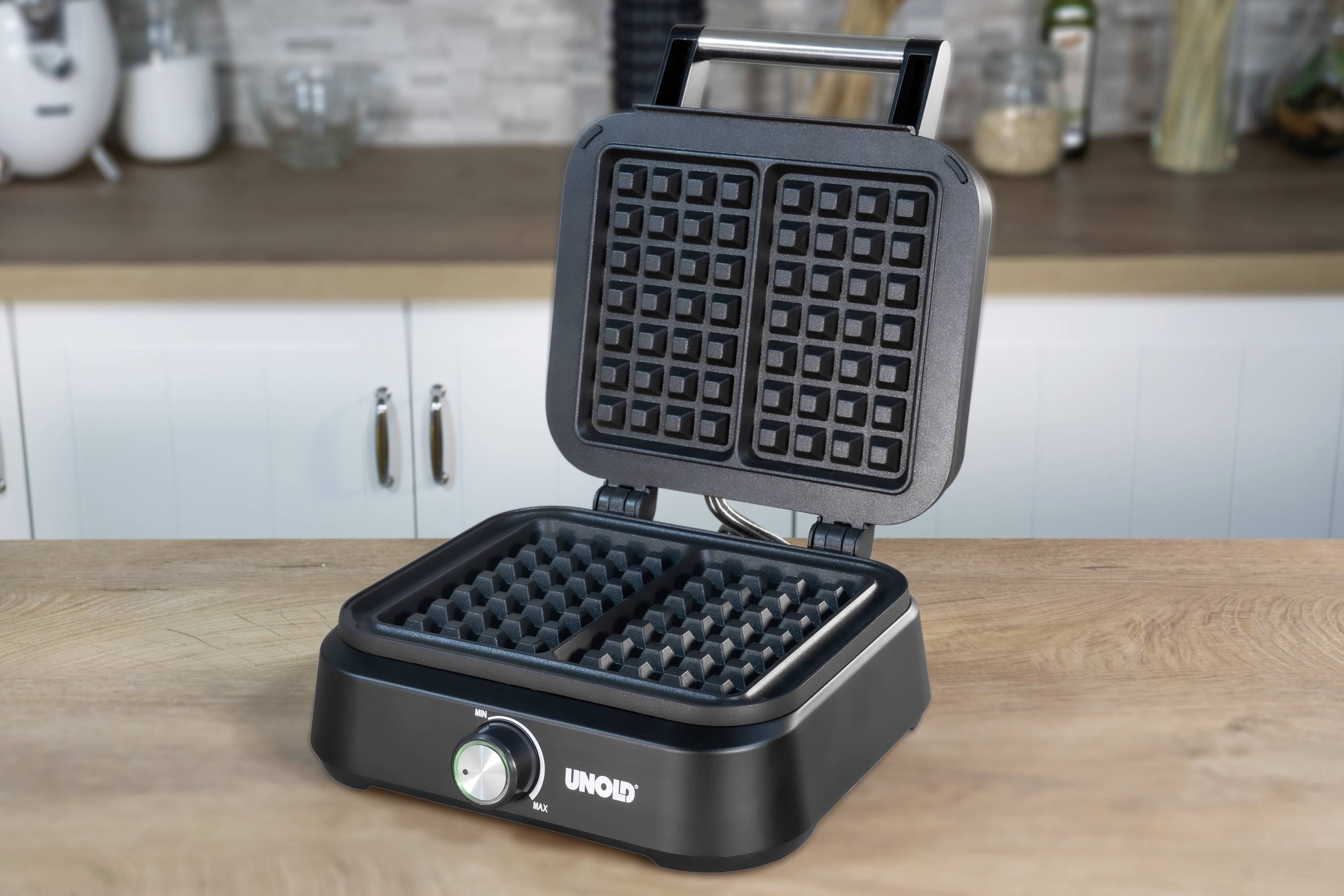 A black waffle maker with an open lid sits on a kitchen worktop. It has a rotary knob for temperature control.