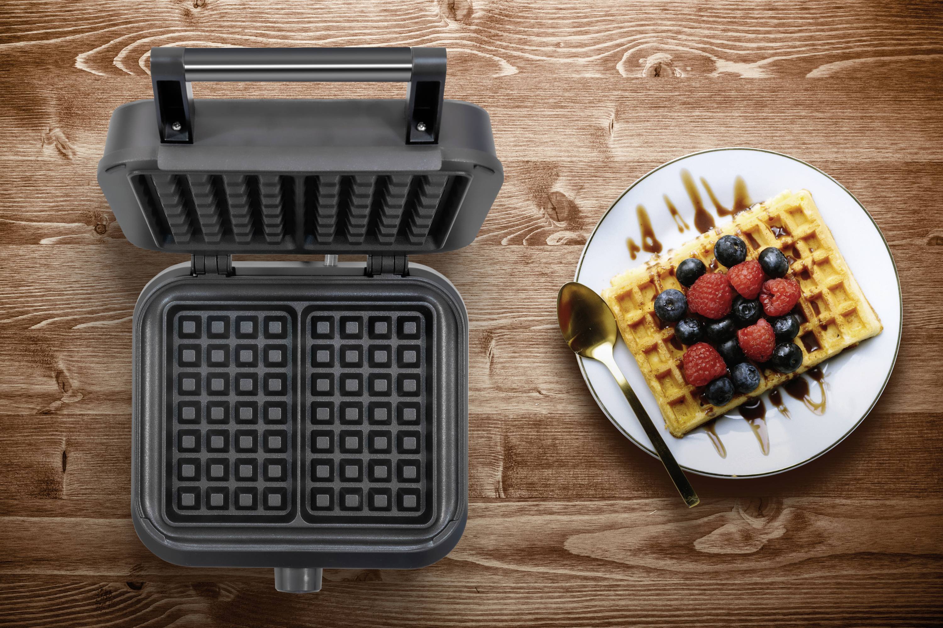 A waffle iron on a wooden surface beside a plate with a waffle garnished with berries and chocolate sauce.