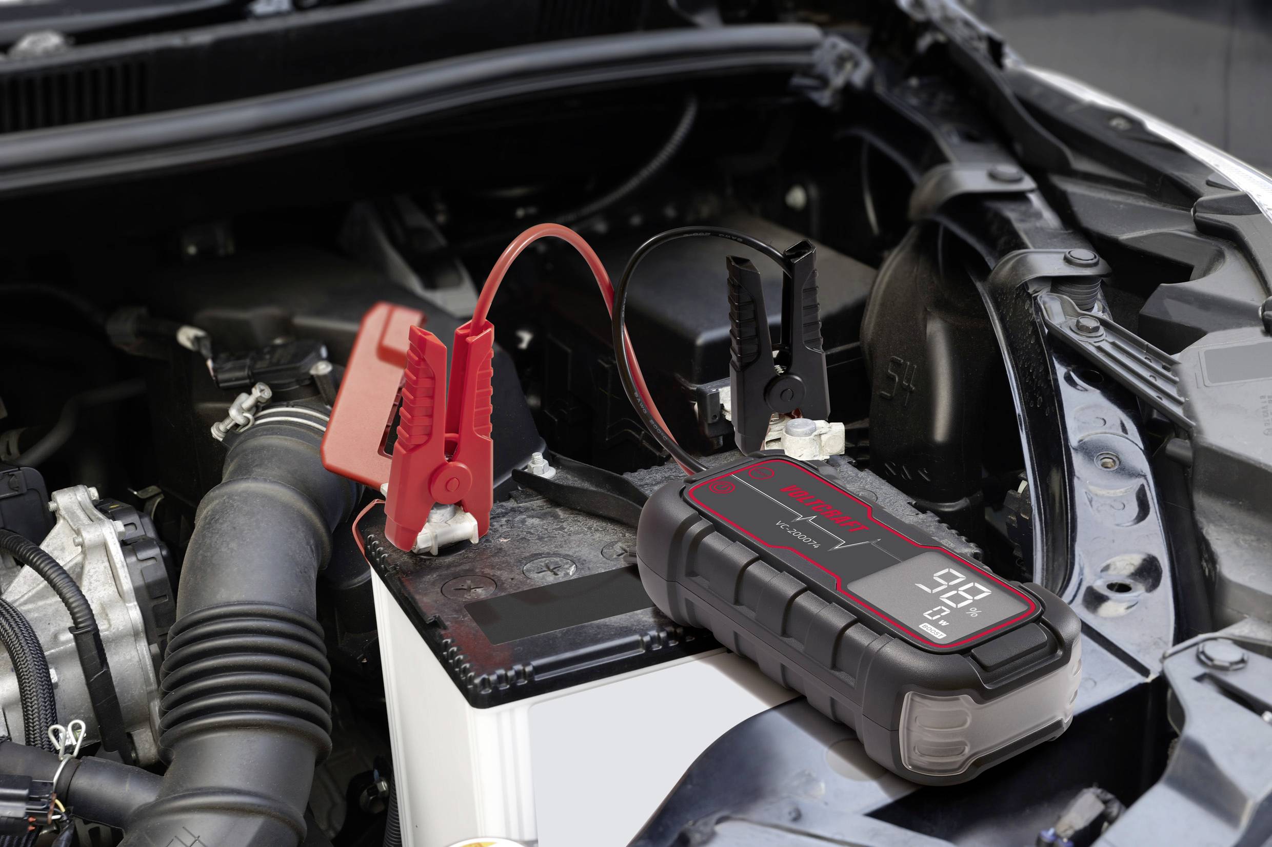 Car battery with connected jump starter, red and black cables attached to battery terminals. Vehicle engine compartment in the background.